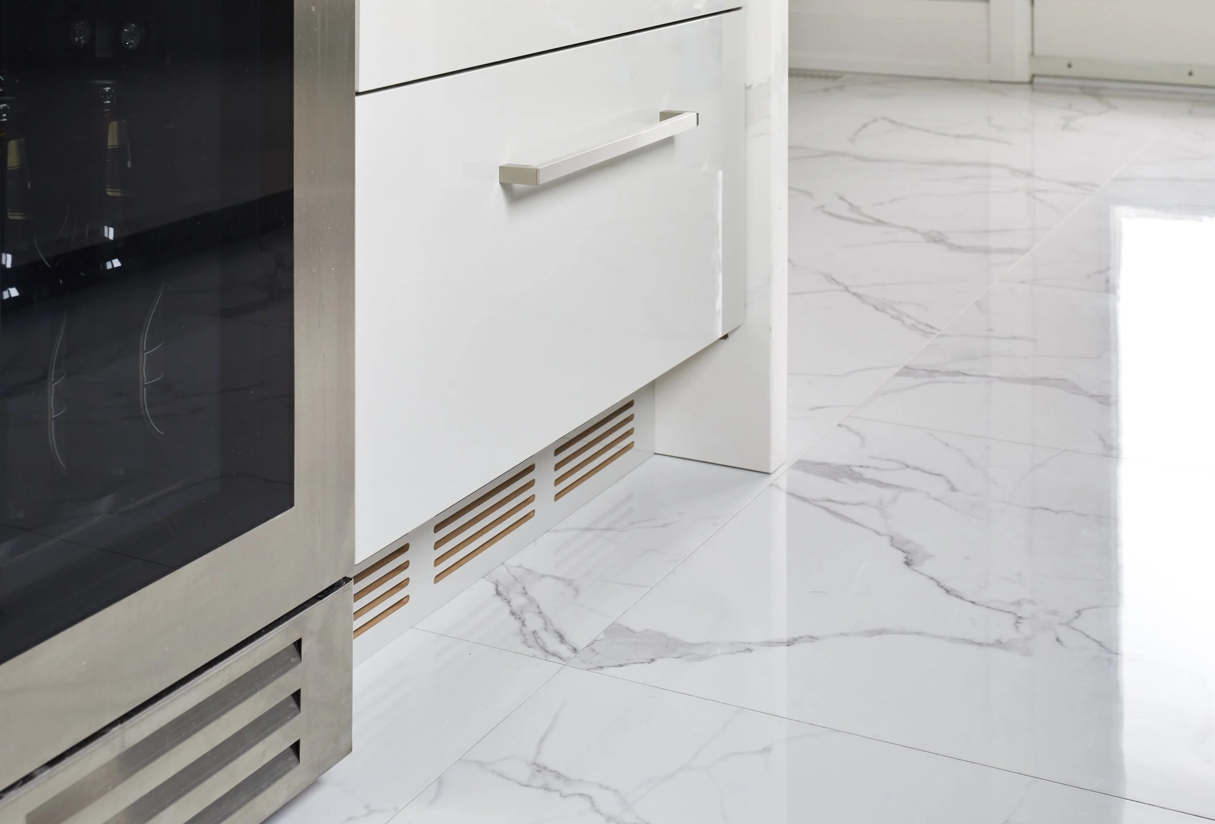 Close-up of a white kitchen cabinet with a metallic handle, a vent below it, and a marble floor with white and gray veining.