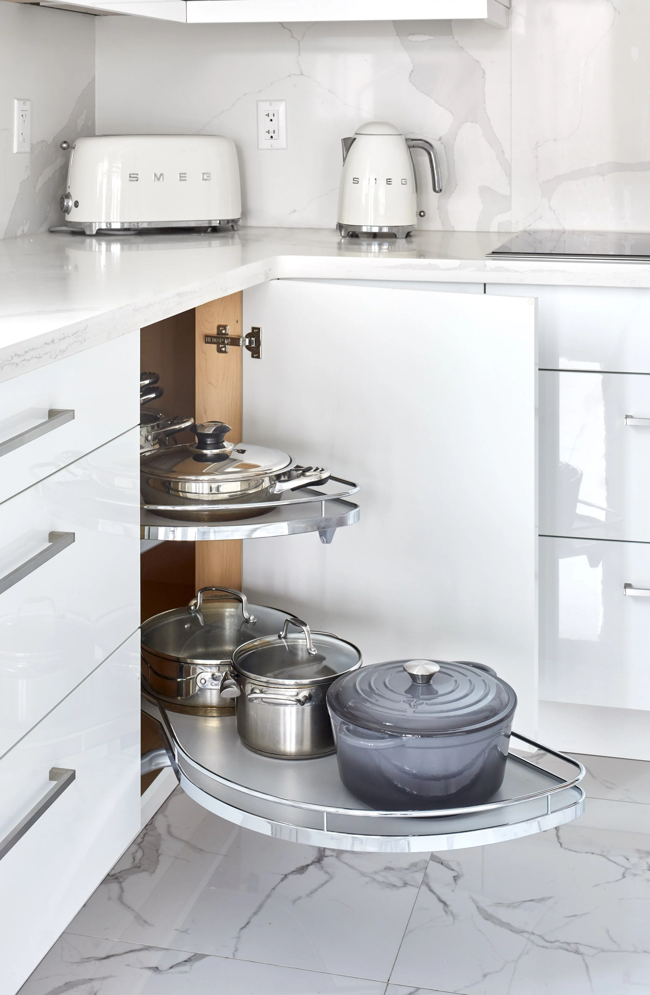 A modern kitchen with white cabinets and marble flooring. The kitchen cabinets are open, revealing pots and pans stored inside. There are electric outlets on the wall, and a small countertop with a toaster, kettle, and cooktop. The open cabinet featu