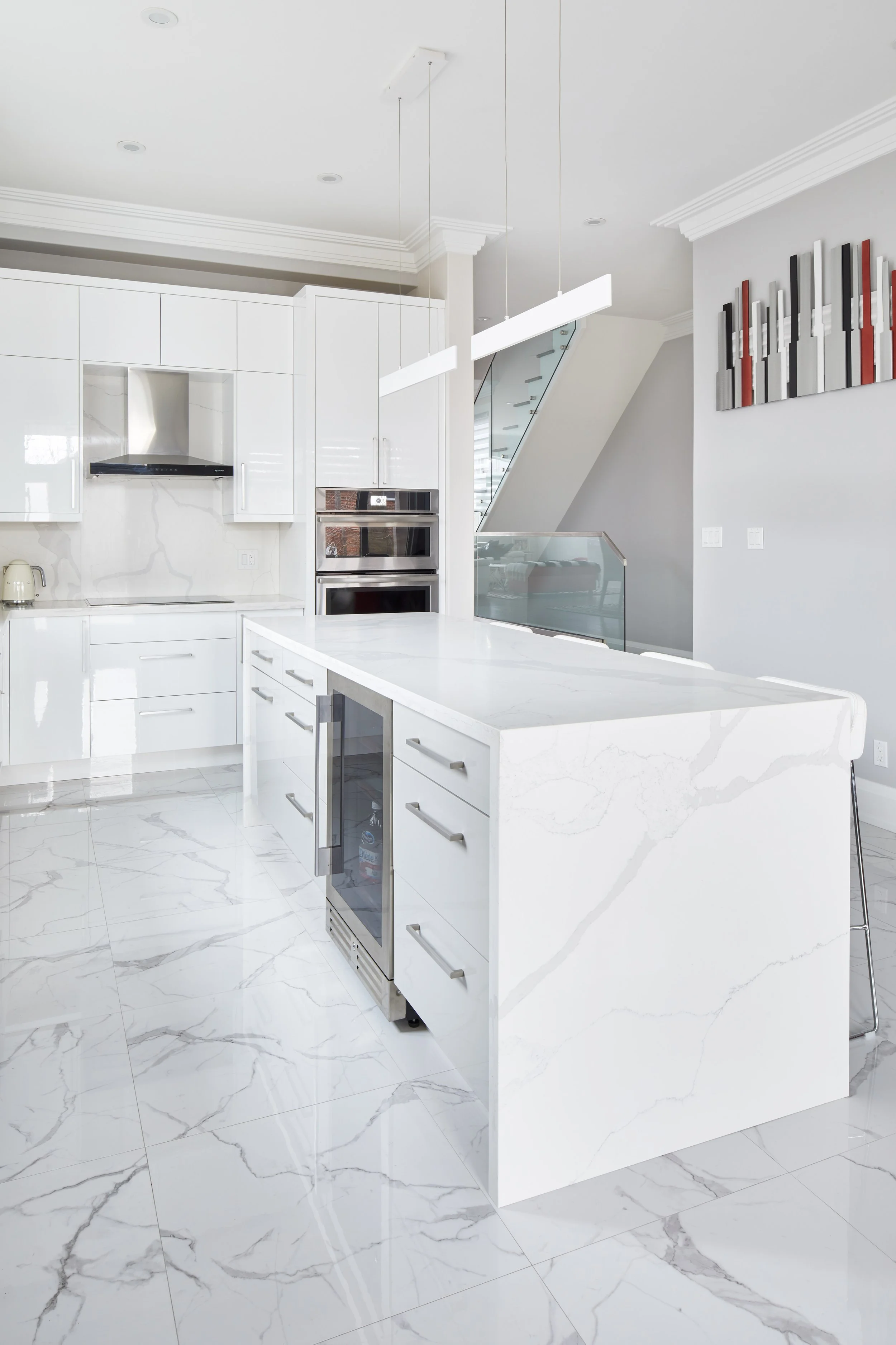 A modern white kitchen with marble countertops and backsplash, white cabinetry, a stainless steel wine cooler, a built-in oven, and artistic wall decor.