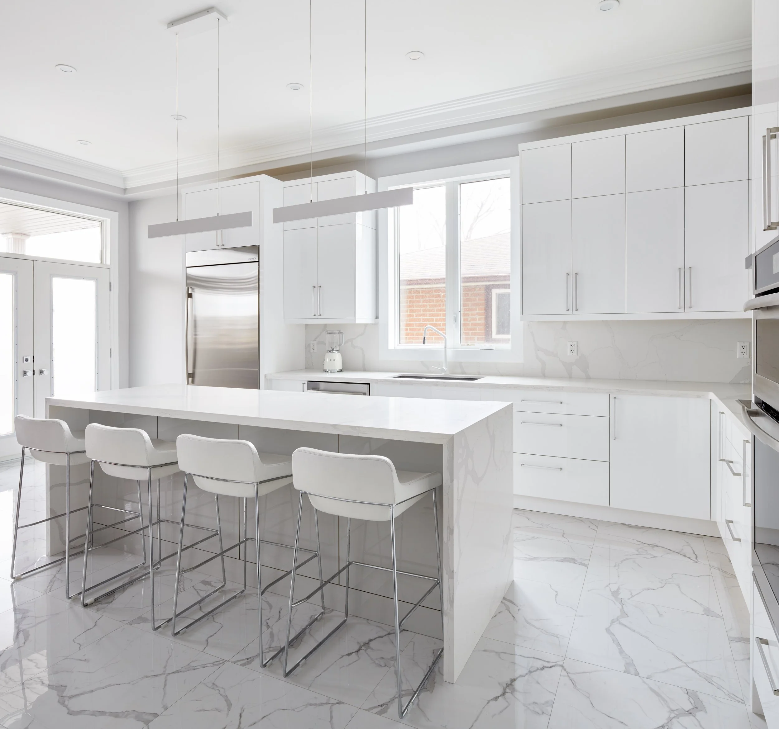 Modern kitchen with white cabinets, marble countertops, and a white island with four white chairs. Large windows and a ceiling light fixture.