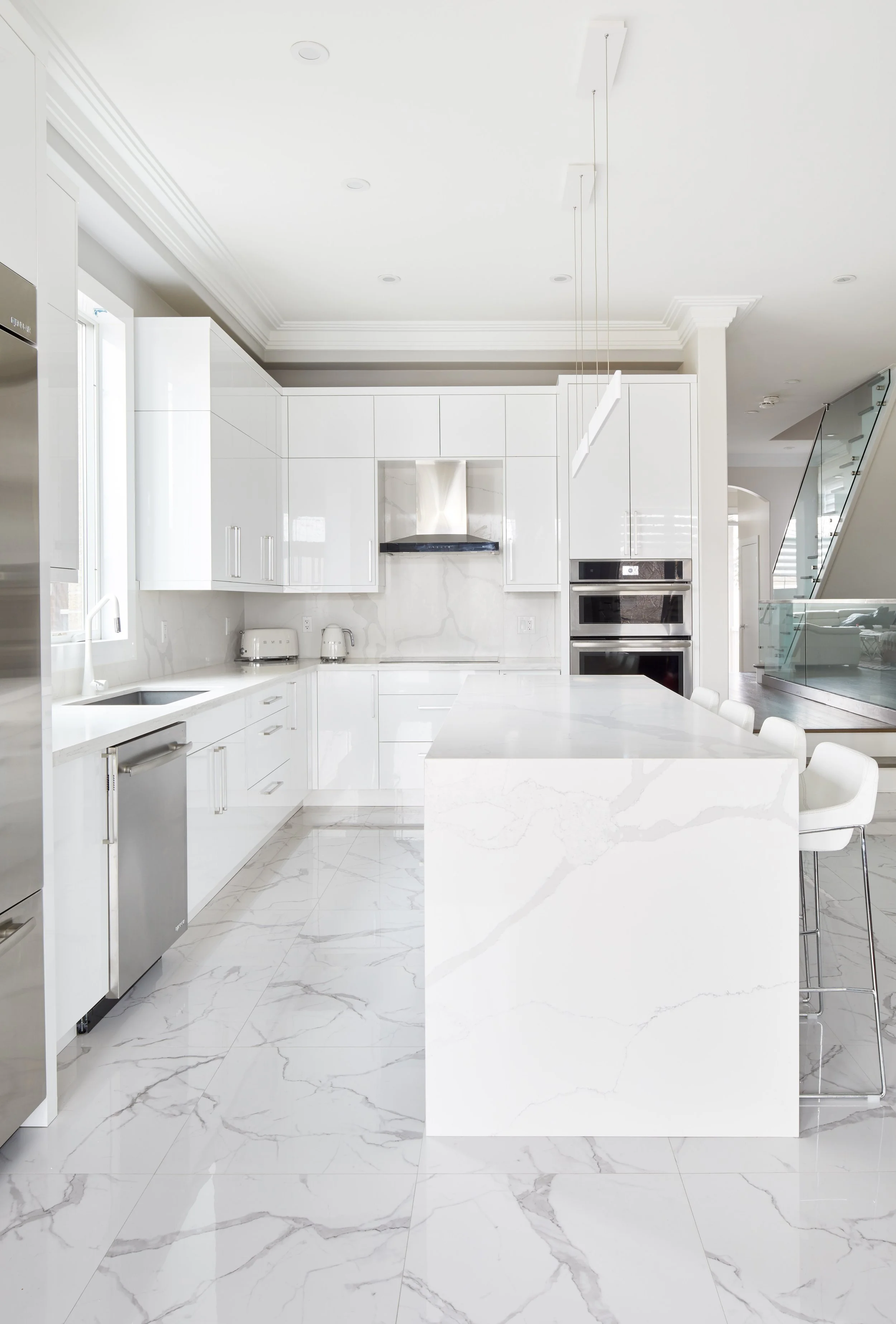 Modern white kitchen with marble countertops and flooring, stainless steel appliances, and a large island with seating.
