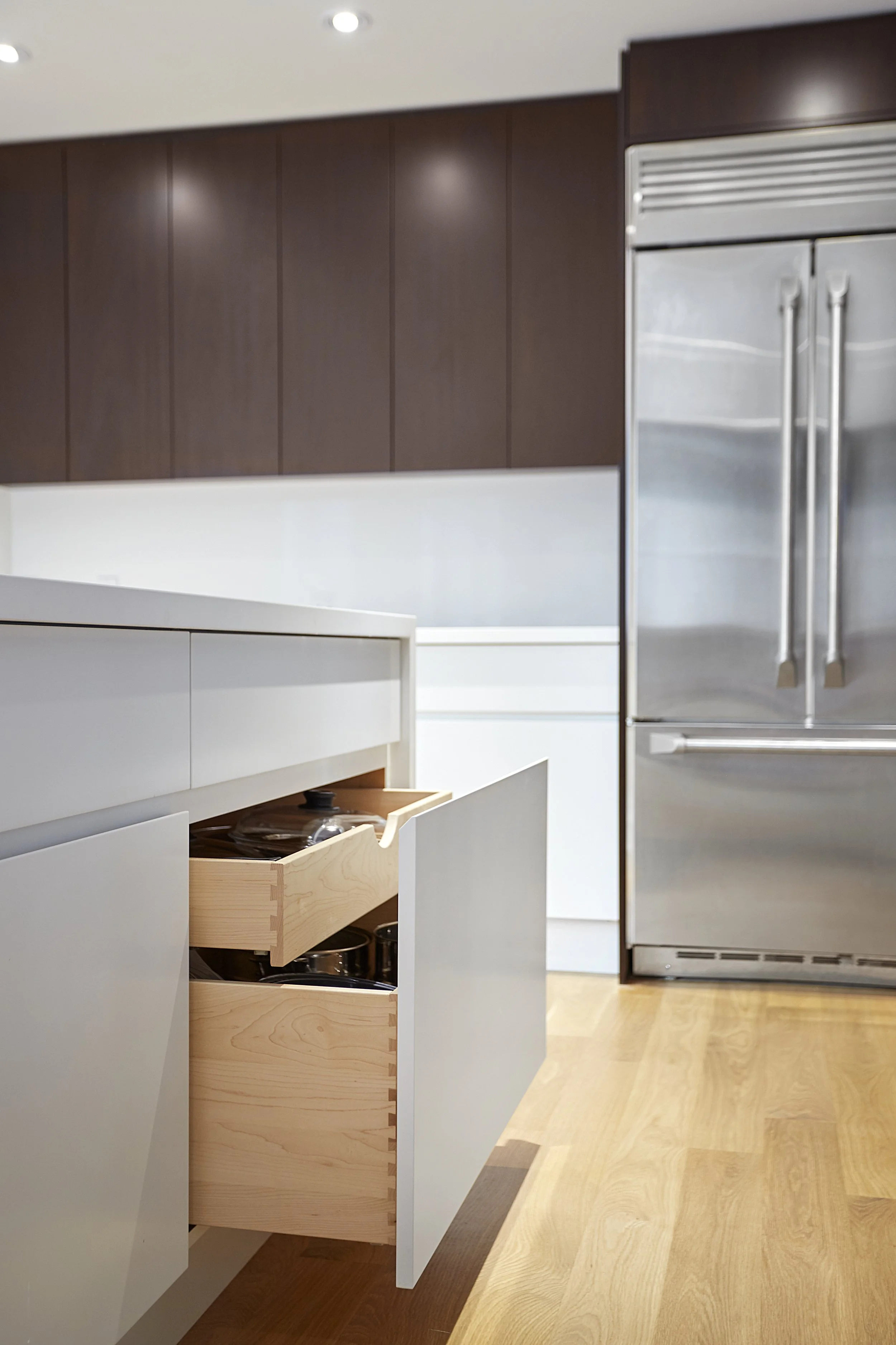 Modern kitchen with dark wood cabinets, white drawers, a stainless steel refrigerator, and wooden flooring.