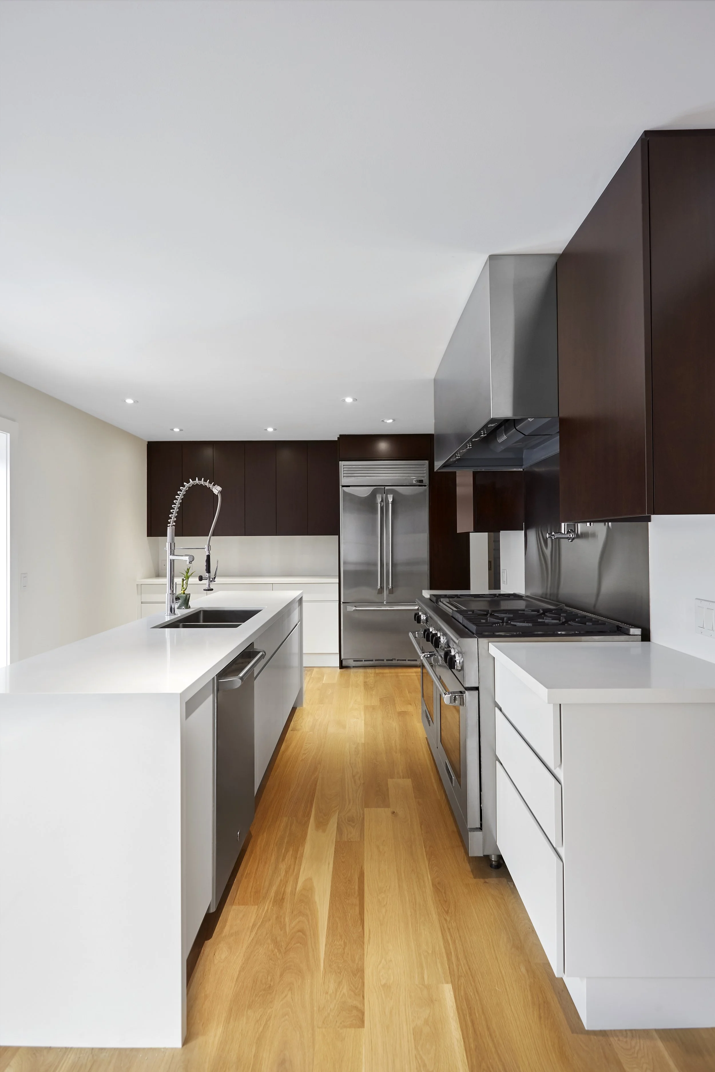Modern kitchen with white countertops, wooden floors, stainless steel appliances, dark wooden cabinets, and a white island with a sink and a gooseneck faucet.