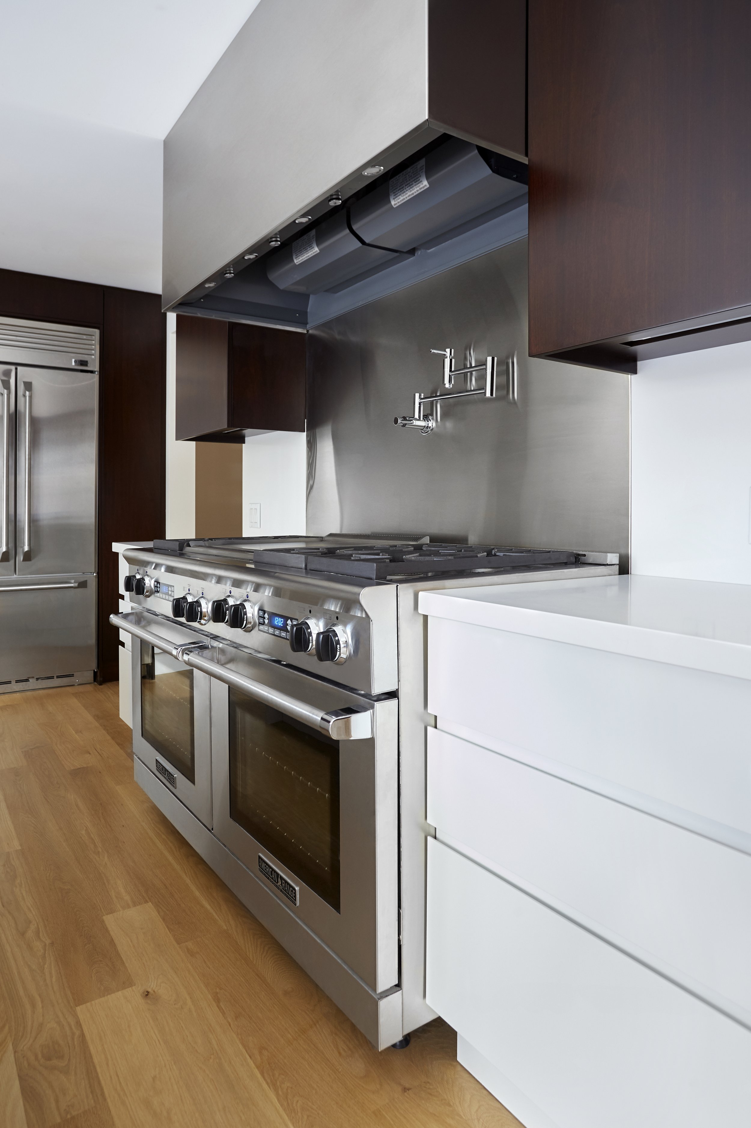 Modern kitchen with stainless steel stove and oven, dark wood cabinets, white drawers, wooden floor, and stainless steel refrigerator.