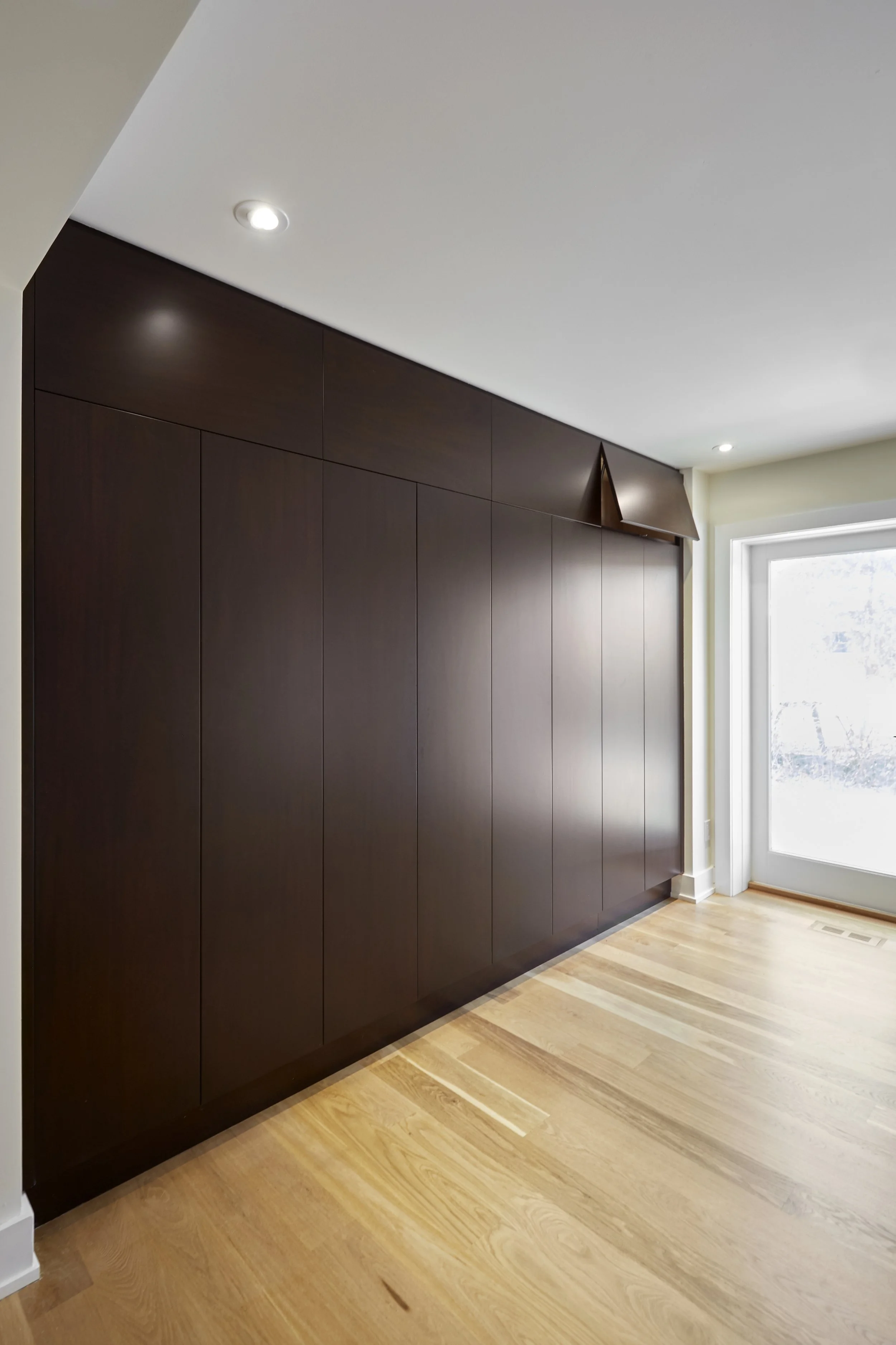A room corner showing a dark wood-paneled wall, hardwood flooring, a window with a white frame, and a small triangular wooden shelf near the ceiling.