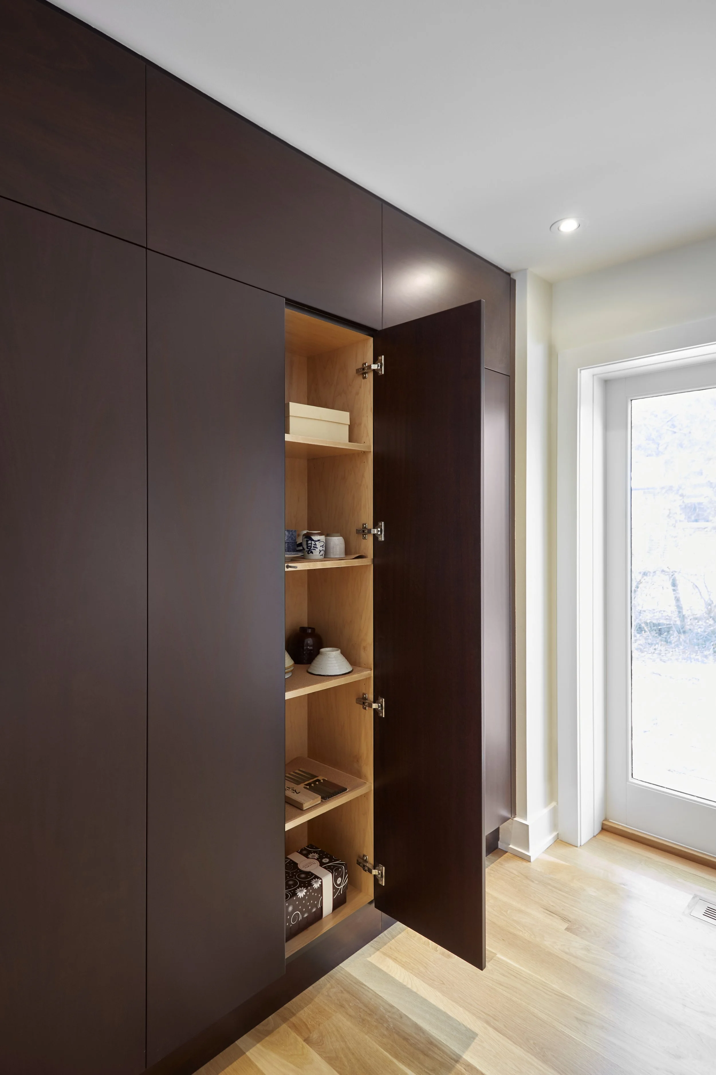 A dark wooden cabinet with an open door revealing shelves with various items near a window in a well-lit room.