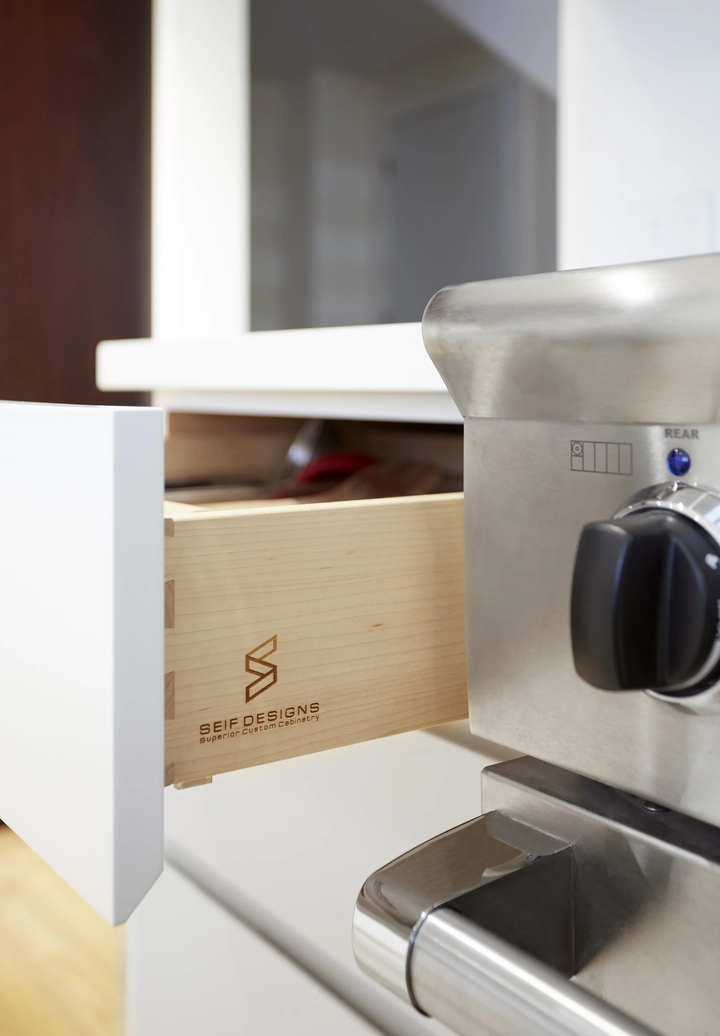 Close-up of a wood drawer with the logo 'SEIF DESIGNS', a silver stove with control knob, and part of a white cabinet with a dark wood finish in the background.