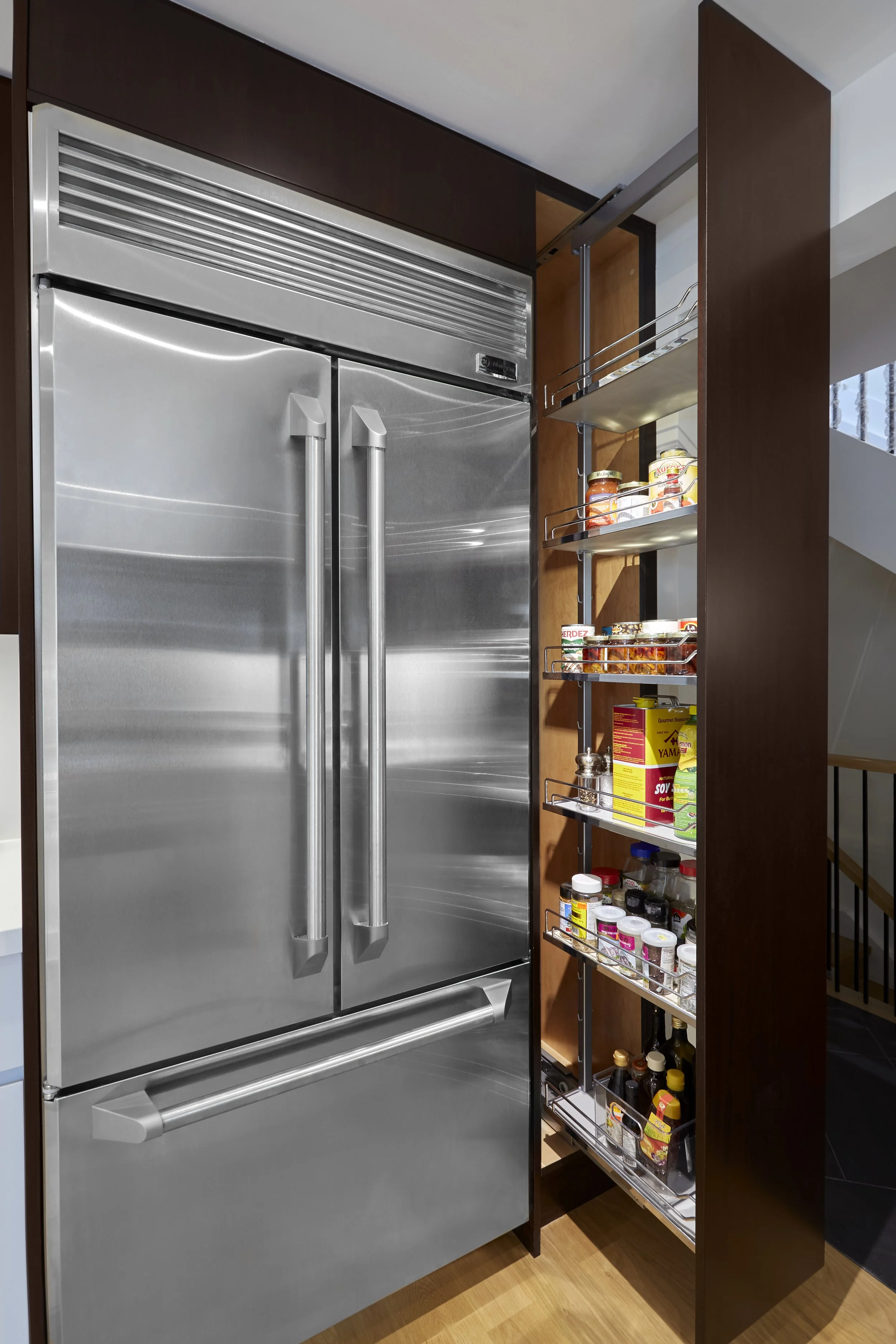 A stainless steel commercial refrigerator with double doors and a dark wood cabinet to the right, featuring a sliding pantry with spices and condiments inside.