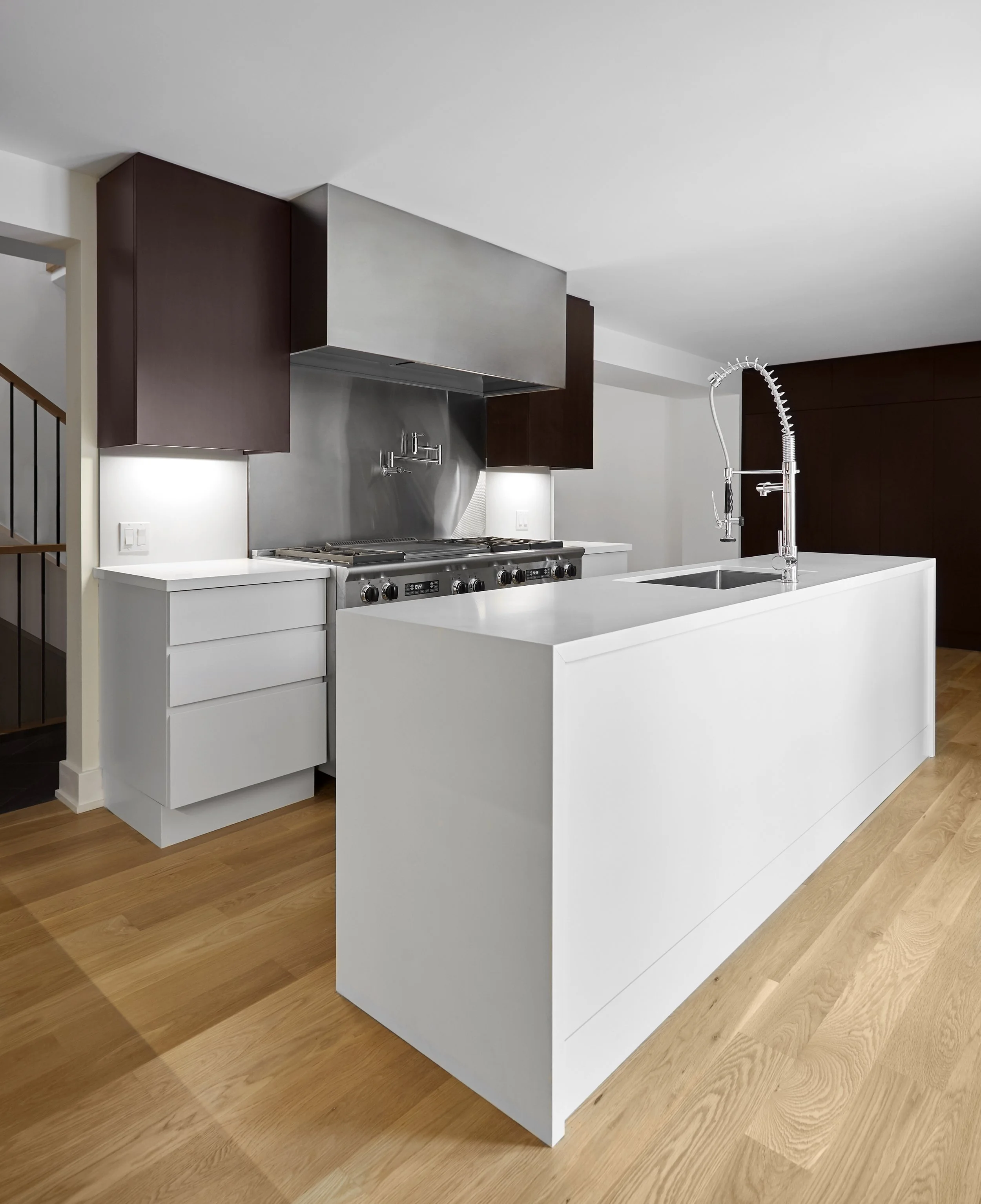 Modern kitchen with white island, dark brown cabinets, stainless steel appliances, and a gold-toned wooden floor.