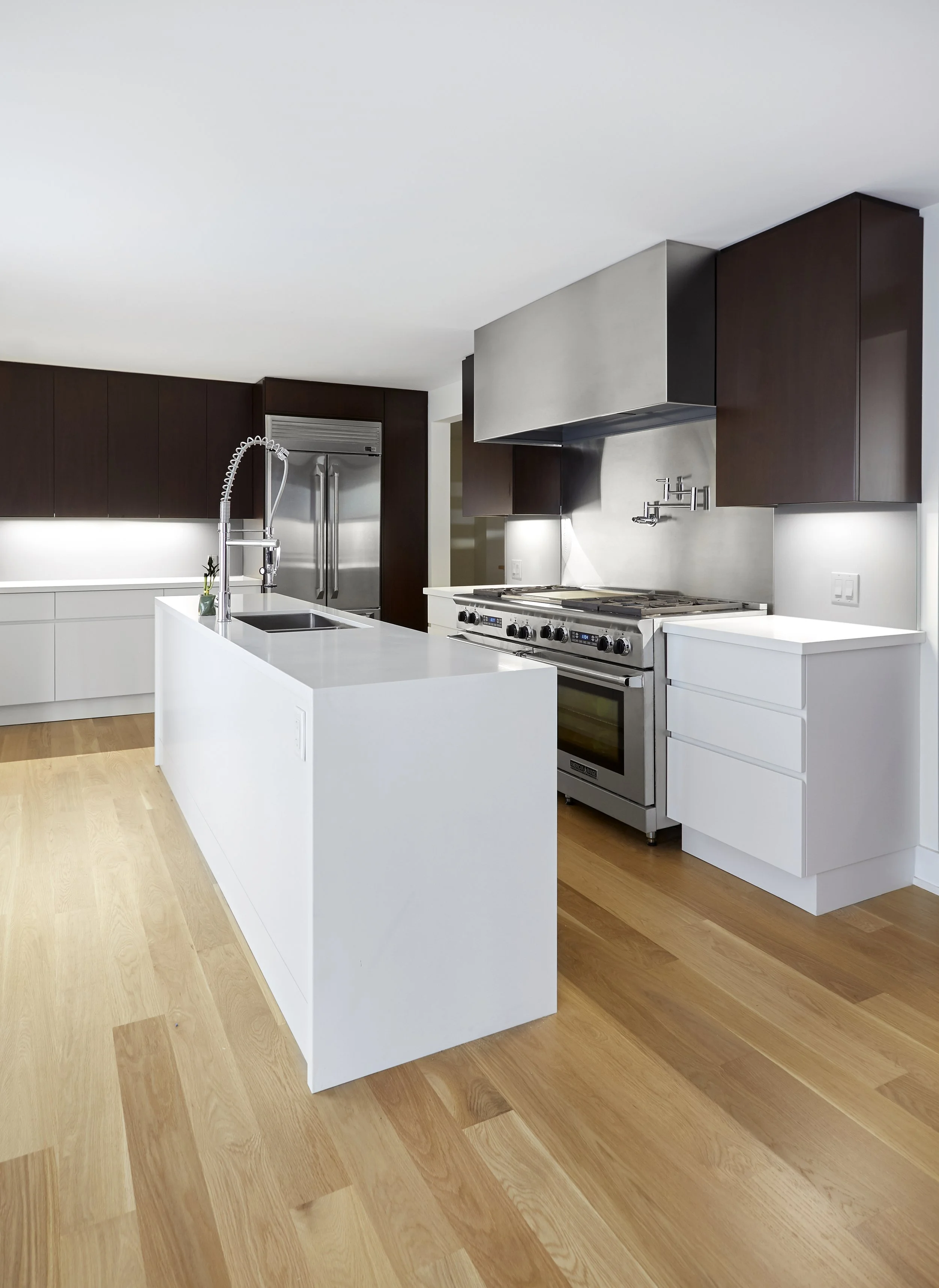 Modern kitchen with white countertops, dark wooden cabinets, stainless steel appliances, and light hardwood flooring.
