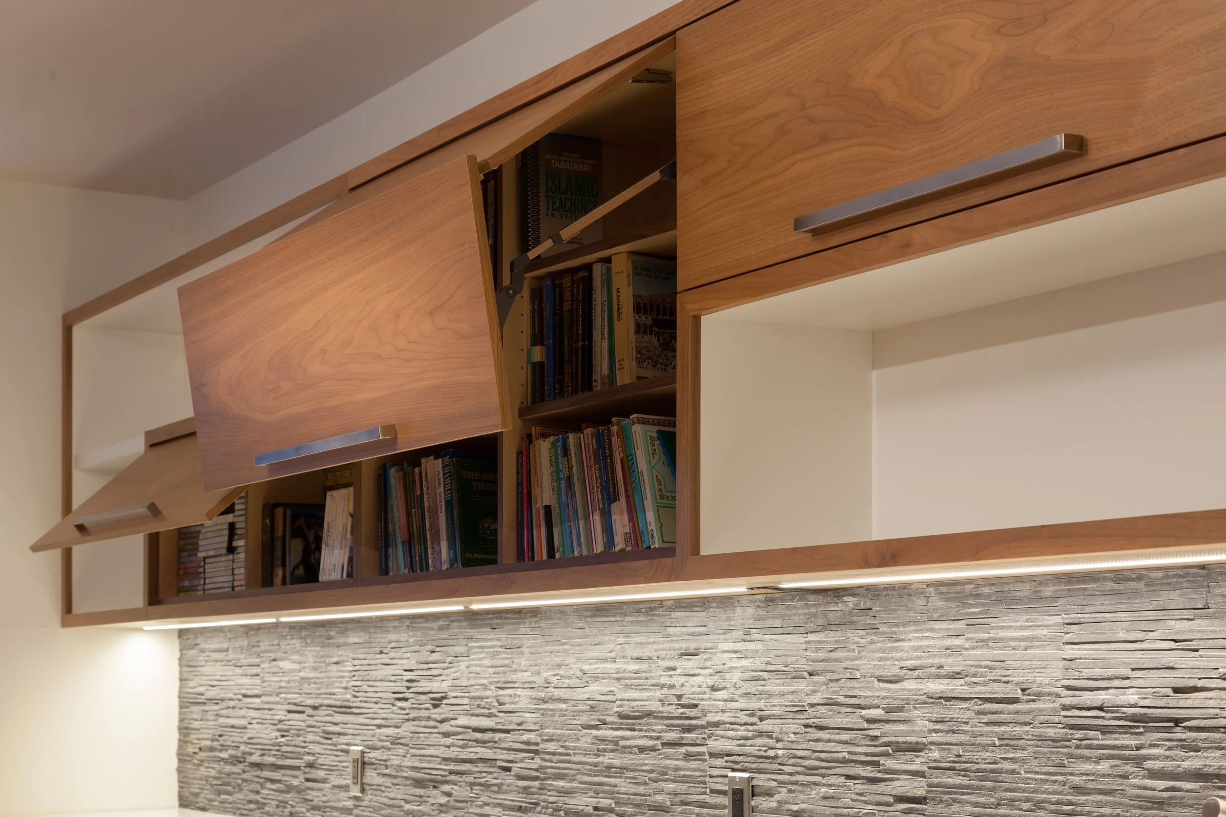 Wooden bookshelf with a tilt-up door, containing books, against a wall with a stone-patterned texture.