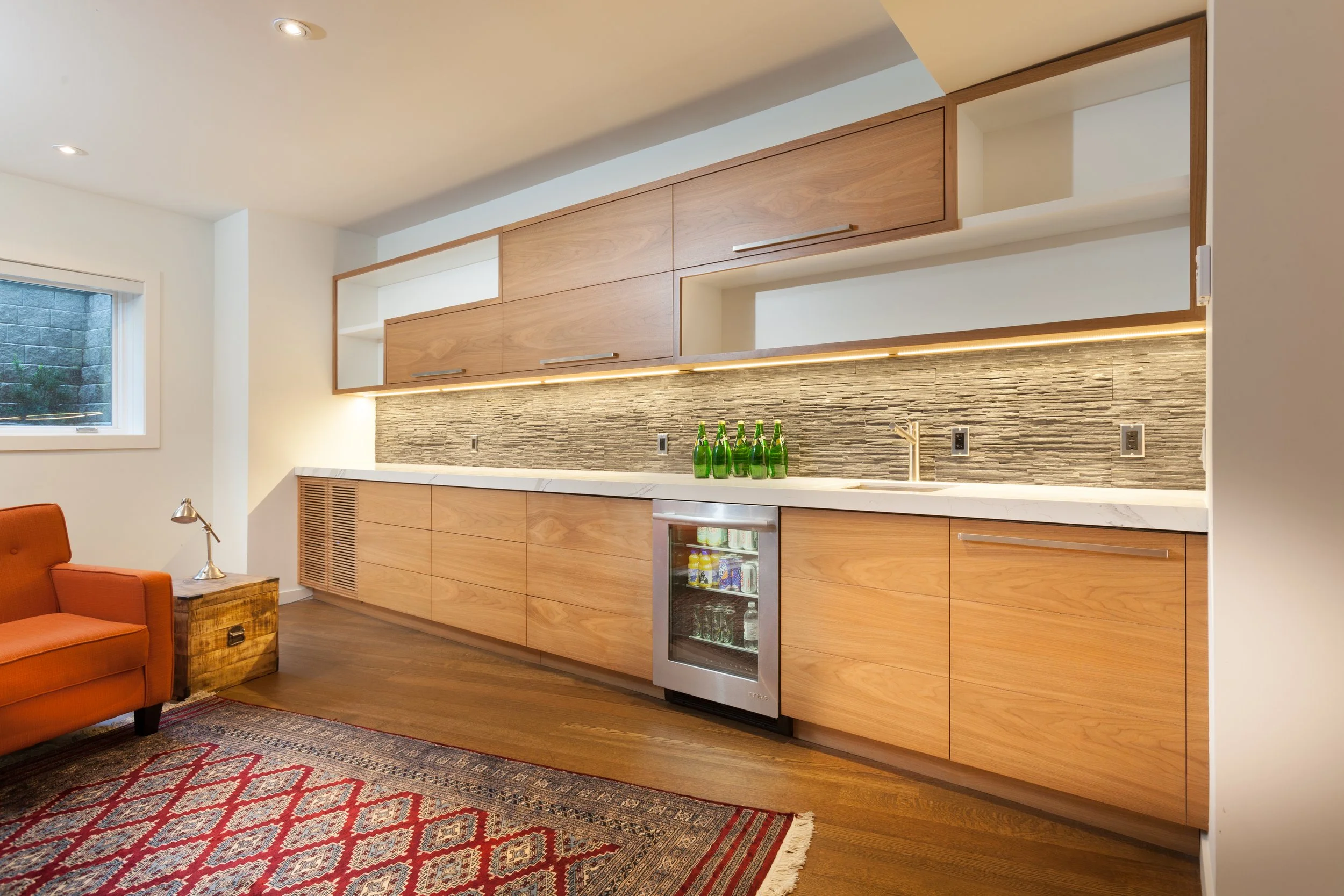 Modern kitchen with wooden cabinets, a textured gray backsplash, a small fridge, and a row of green bottles on the countertop. Part of a living area with an orange armchair and a wooden side table.