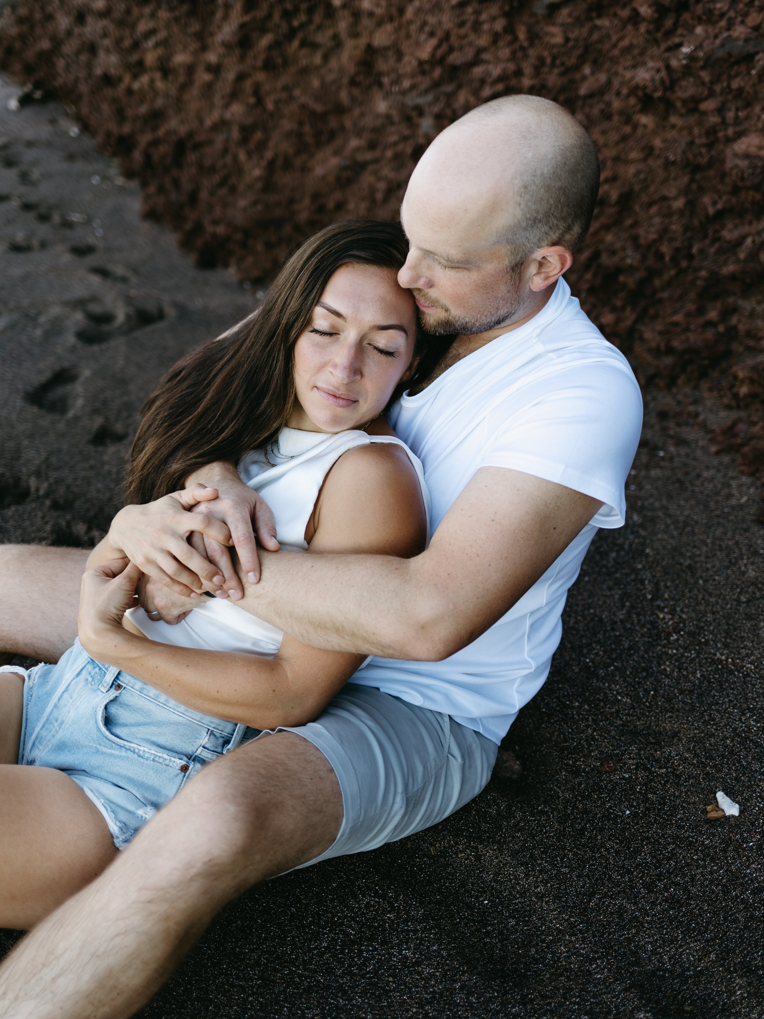 Couple embracing on a dark sandy beach, woman in denim shorts, man in white shirt and shorts.