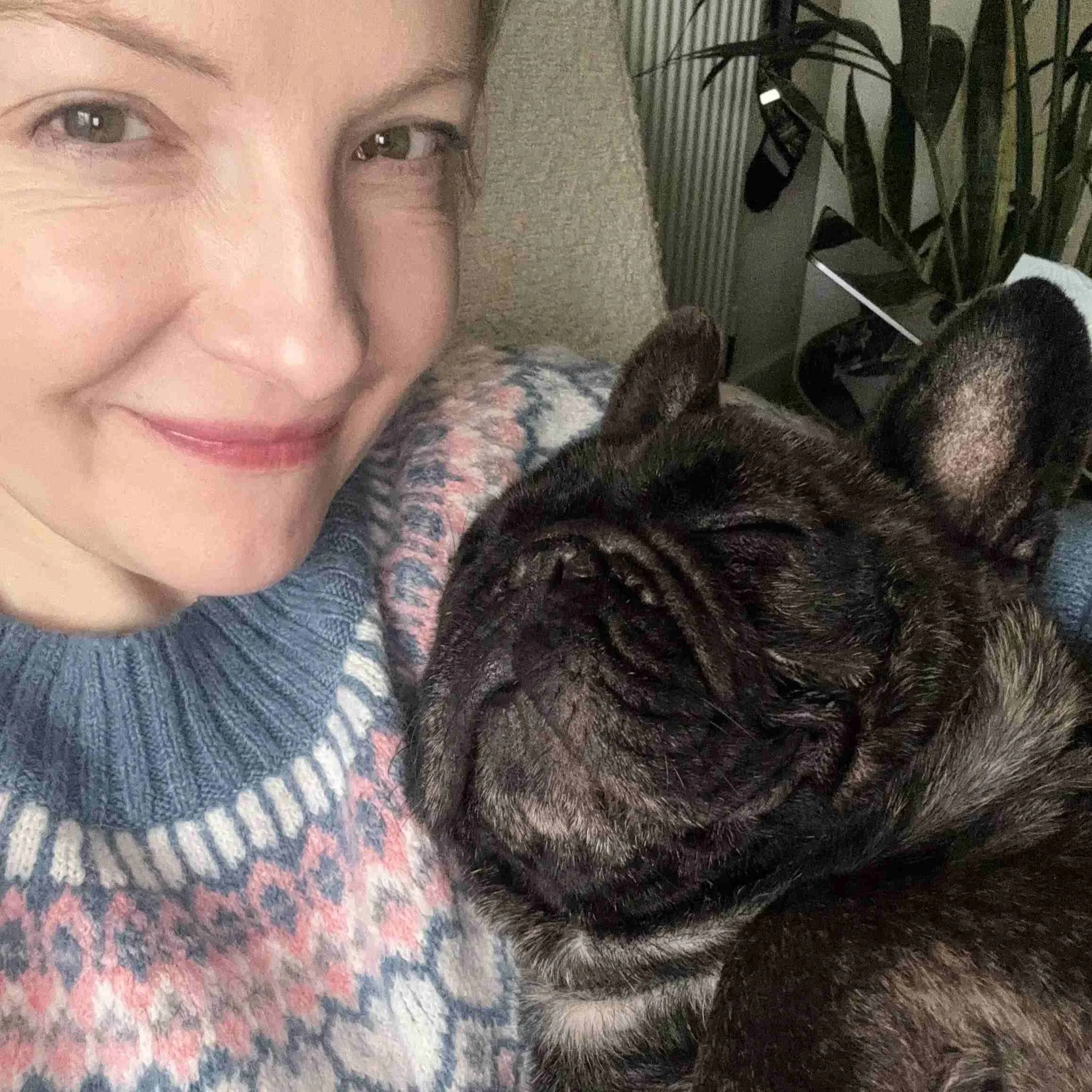 A woman with short blonde hair smiling at the camera while snuggling with a dark brindle French Bulldog resting its head on her shoulder.