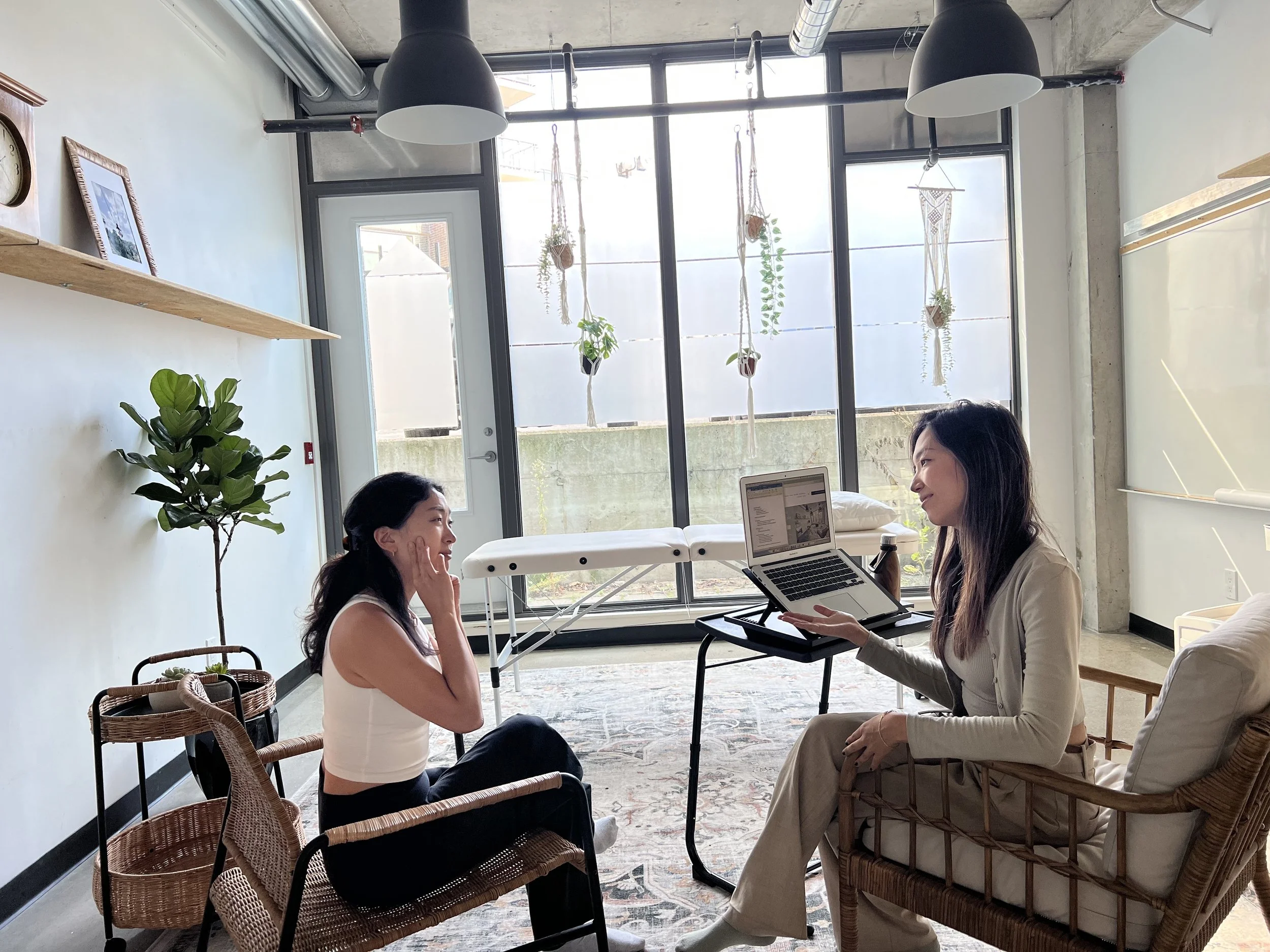 Two women sitting in a cozy room, one using a laptop and the other touching her face. The room has large windows, plants, hanging decorations, and wooden shelves. They are seated in wicker chairs.