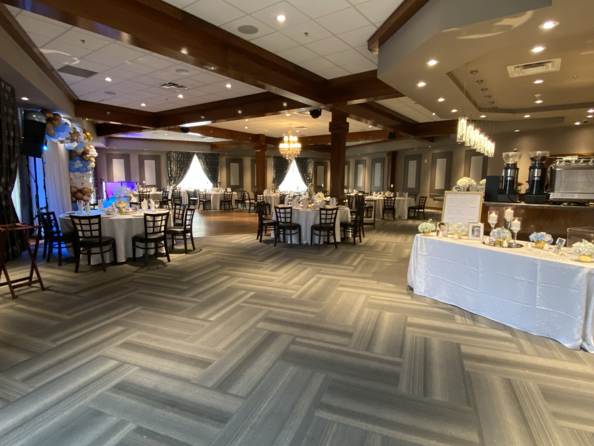 An elegantly decorated banquet hall with multiple round tables covered with white tablecloths, surrounded by wooden chairs, and a dessert or display table on the right side with framed photos, flowers, and candles. The hall features large windows wit