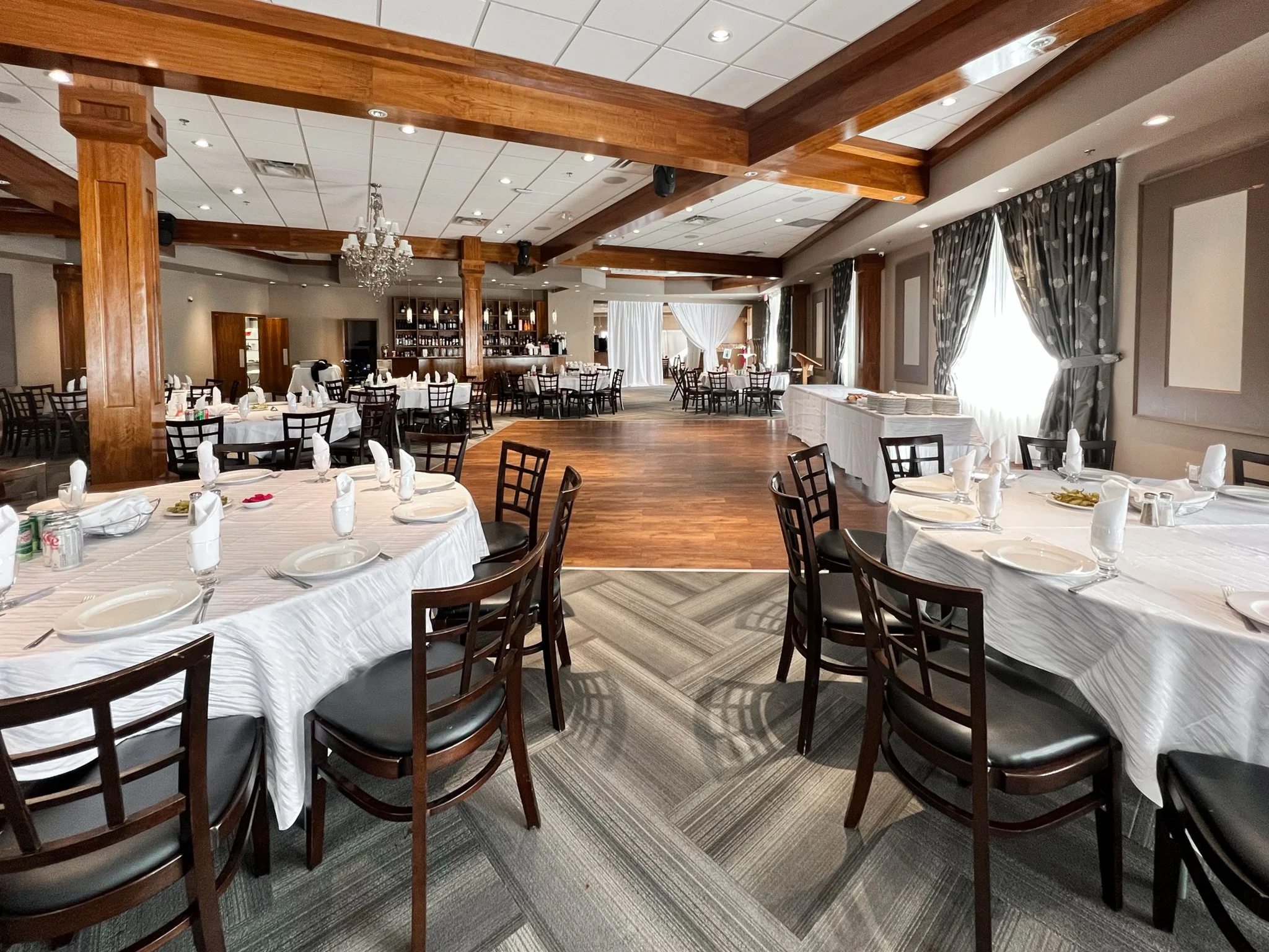 An elegantly decorated banquet hall with round tables covered in white tablecloths, set with plates, glasses, silverware, and folded napkins. The room features wooden columns, a chandelier, and large windows with patterned curtains, with a wooden dance floor in the center and a bar area in the background.