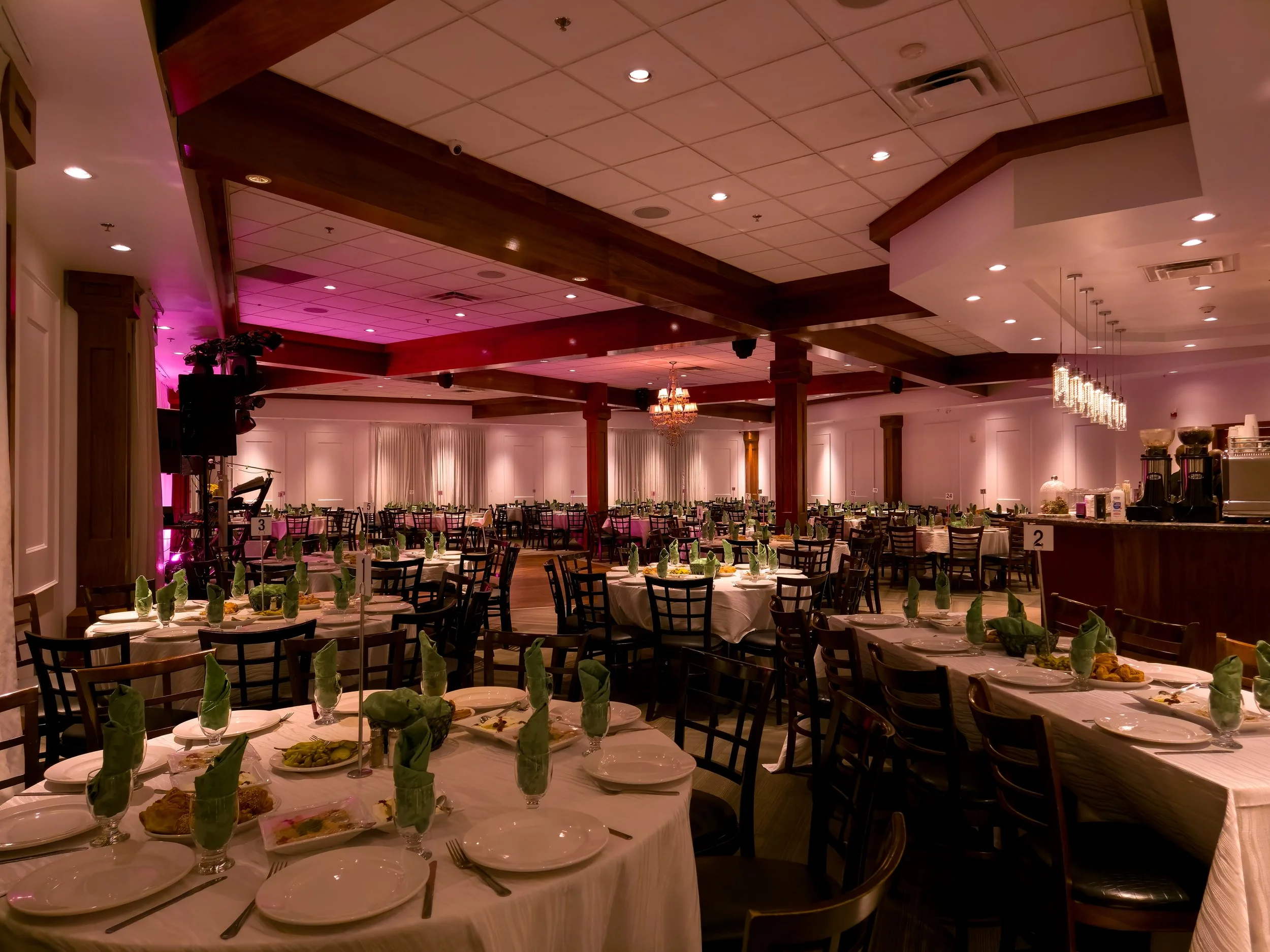 Banquet hall decorated for an event with round tables covered with white tablecloths, set with plates, utensils, glasses with green napkins, and food. There are chairs around the tables, a buffet area on the right with coffee machines and containers,