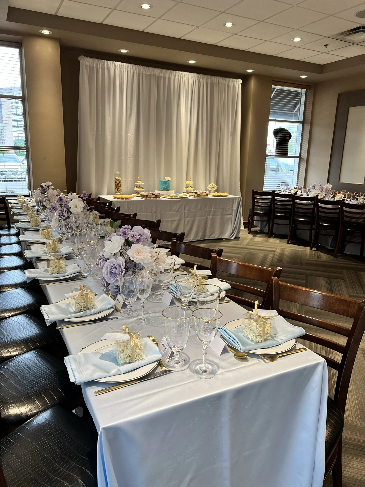 Decorated banquet table set for a celebration with purple and white flowers, gold utensils, and small gift boxes, in a room with windows and a dessert table in the background.