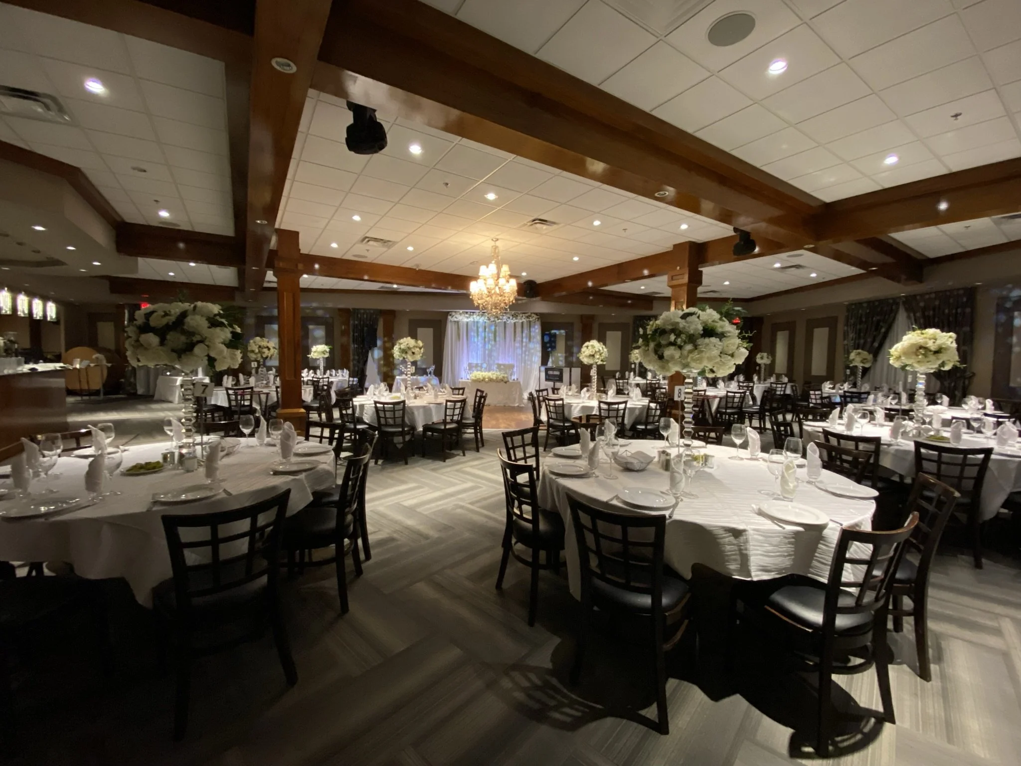 An elegantly decorated banquet hall with round tables covered in white tablecloths, each adorned with a large floral centerpiece of white flowers. The room features wooden beams on the ceiling, a chandelier, and large windows with dark curtains, crea