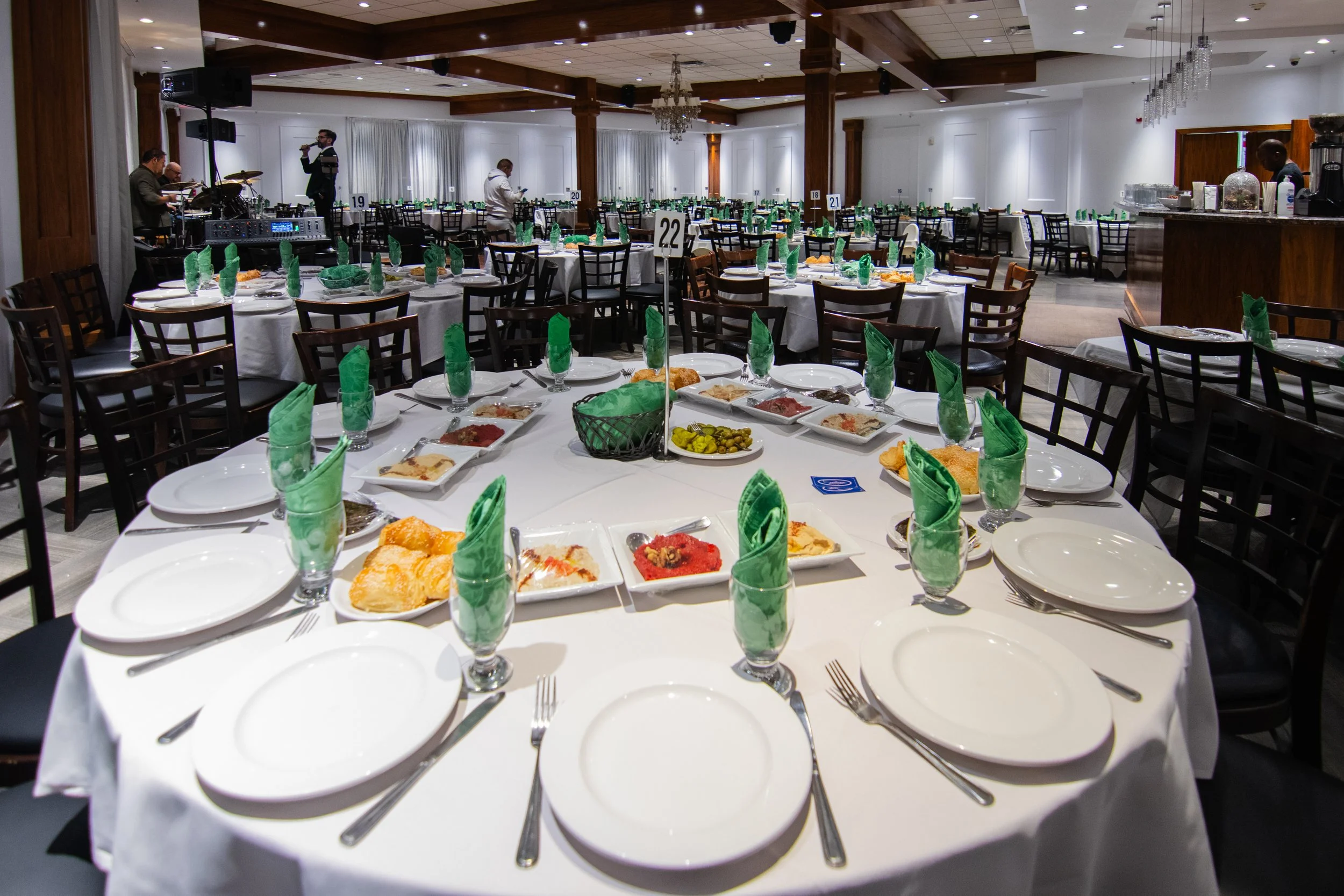 A banquet hall set up for a formal event with round tables covered in white tablecloths, green napkins folded in glasses, and plates with food. There is a stage with musical instruments and people preparing in the background.