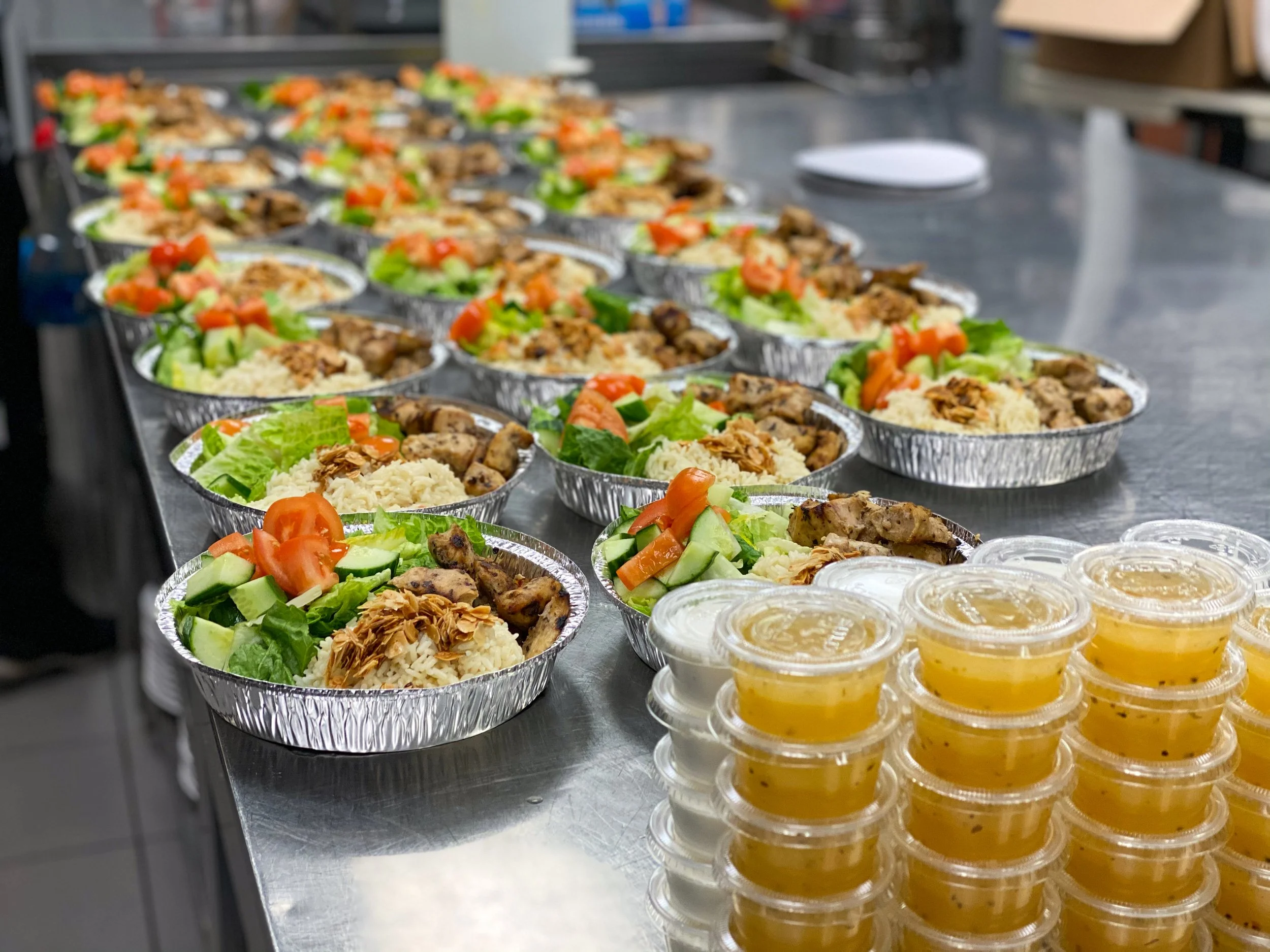 Multiple aluminum food containers filled with rice, grilled chicken, fresh vegetables such as lettuce, cucumbers, and tomatoes, arranged on a stainless steel table, with small plastic containers of yellow sauce in front.