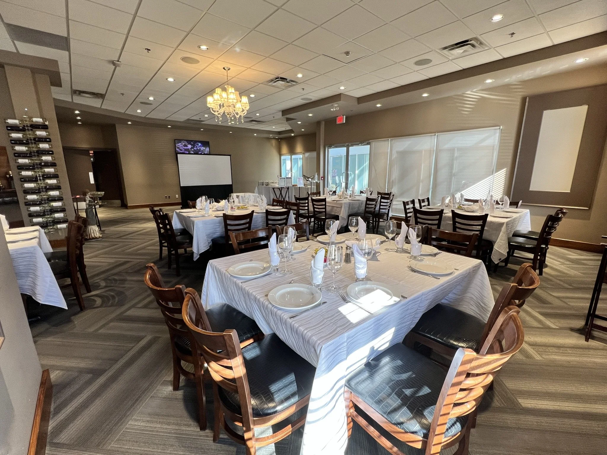 Restaurant dining room with set tables, white tablecloths, wine glasses, plates, and utensils, with sunlight streaming through windows.
