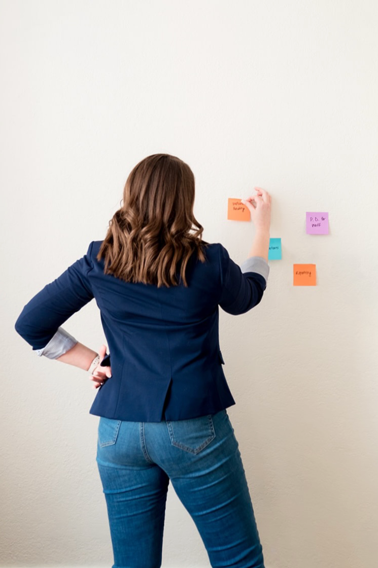 A woman in a blue blazer and jeans standing in front of a wall with colorful sticky notes, rearranging or reviewing the notes.