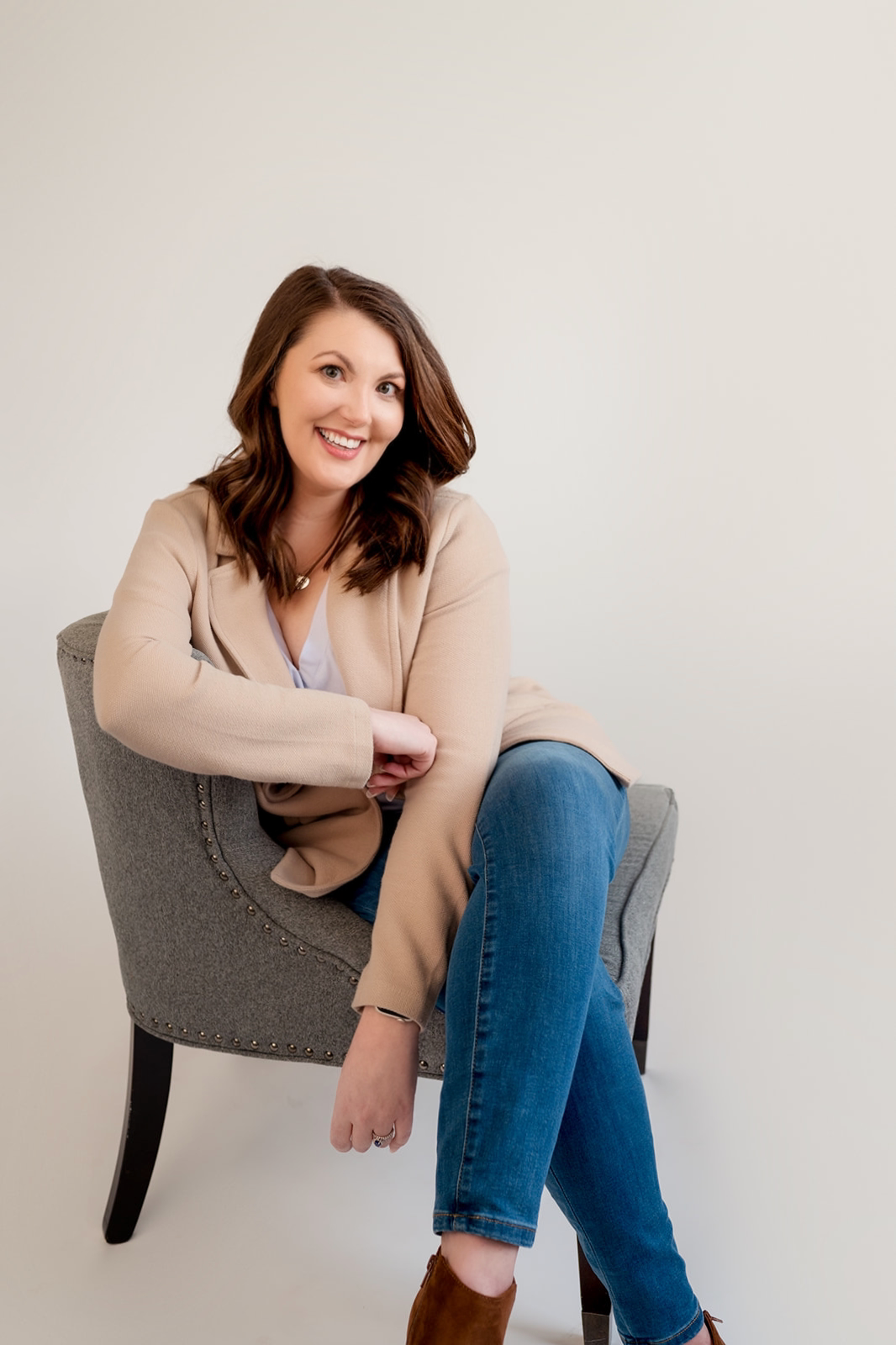 A smiling woman with shoulder-length brown hair, wearing a beige blazer, white top, blue jeans, and brown boots, sitting in a gray upholstered chair against a plain white background.