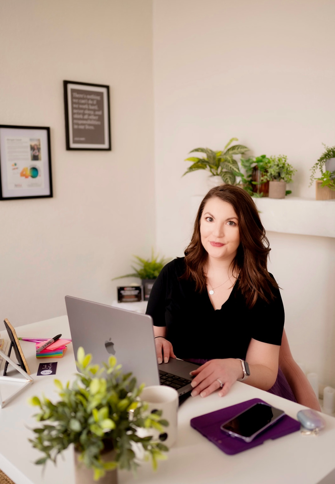 A woman with shoulder-length brown hair, wearing a black top, sitting at a desk with a laptop
