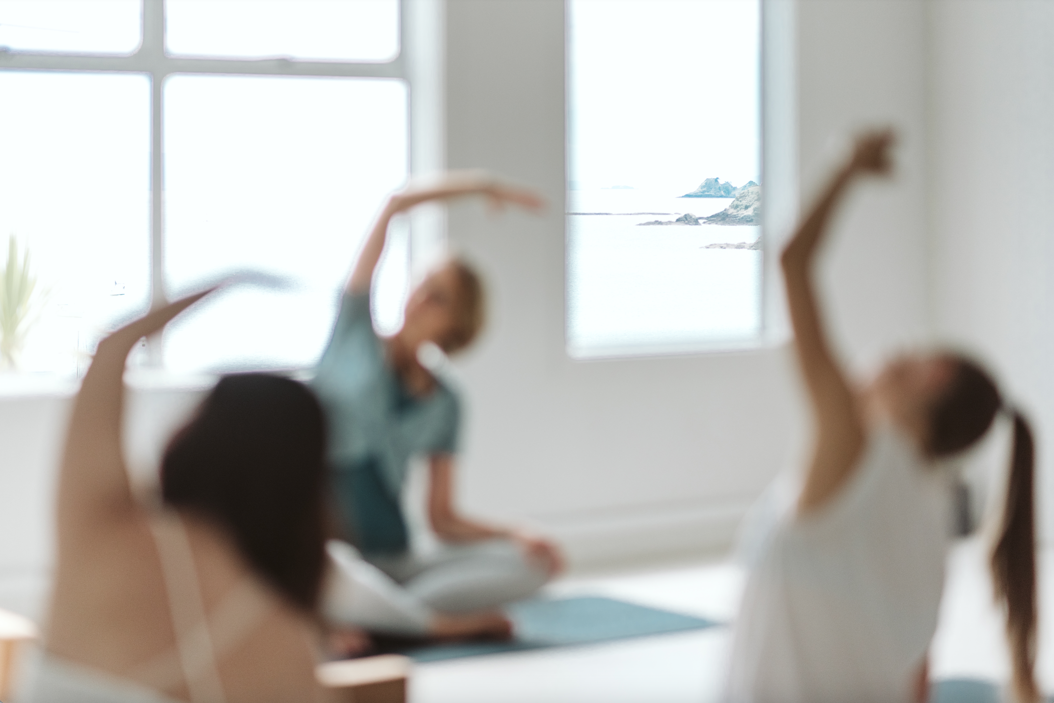 Three people practicing yoga in a bright room with large windows showing a scenic ocean view.