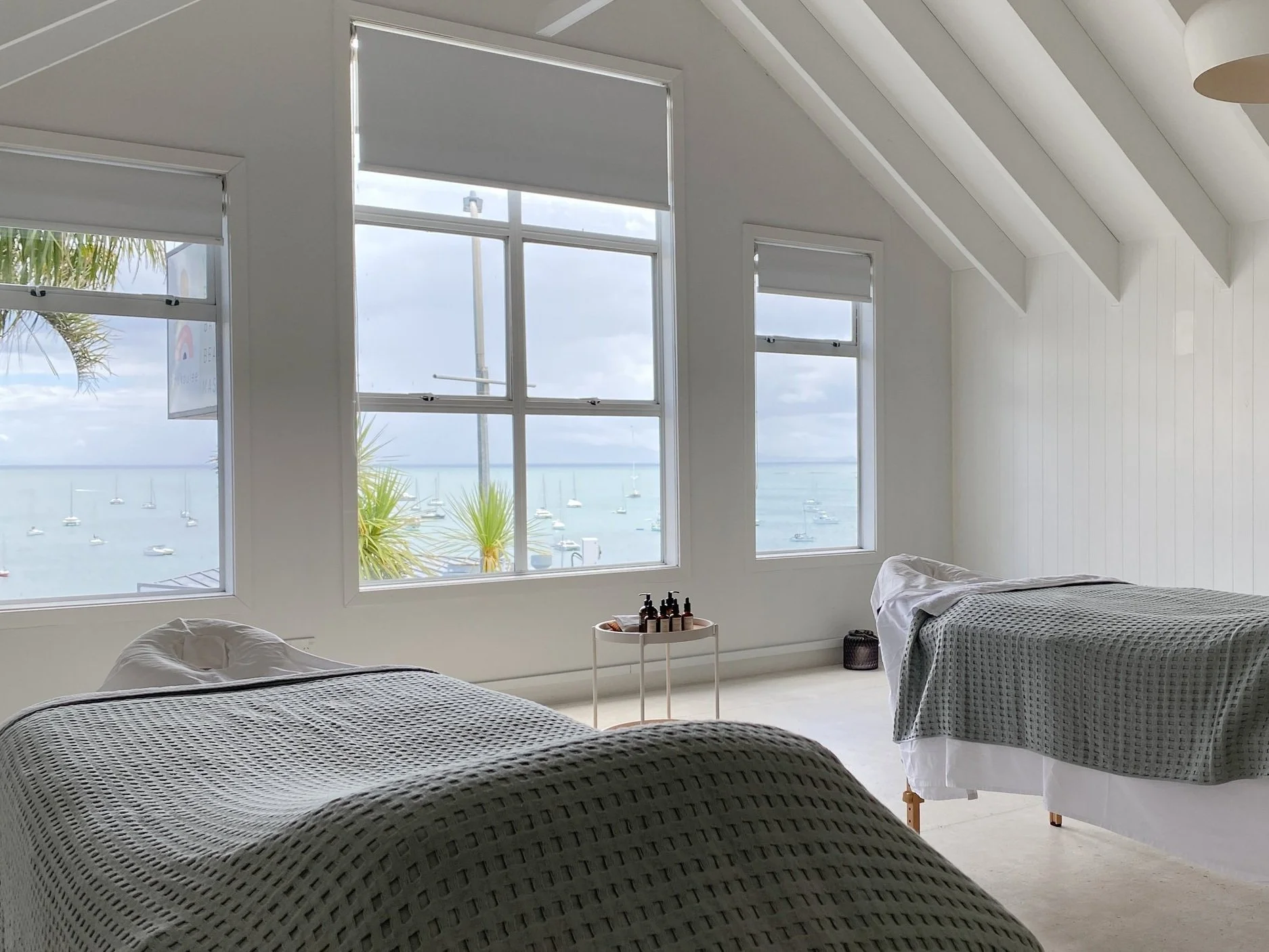 Interior of a spa or massage room with two massage tables covered with gray blankets, large windows showing a view of the ocean with boats, and a small table with bottles of oils or lotions.
