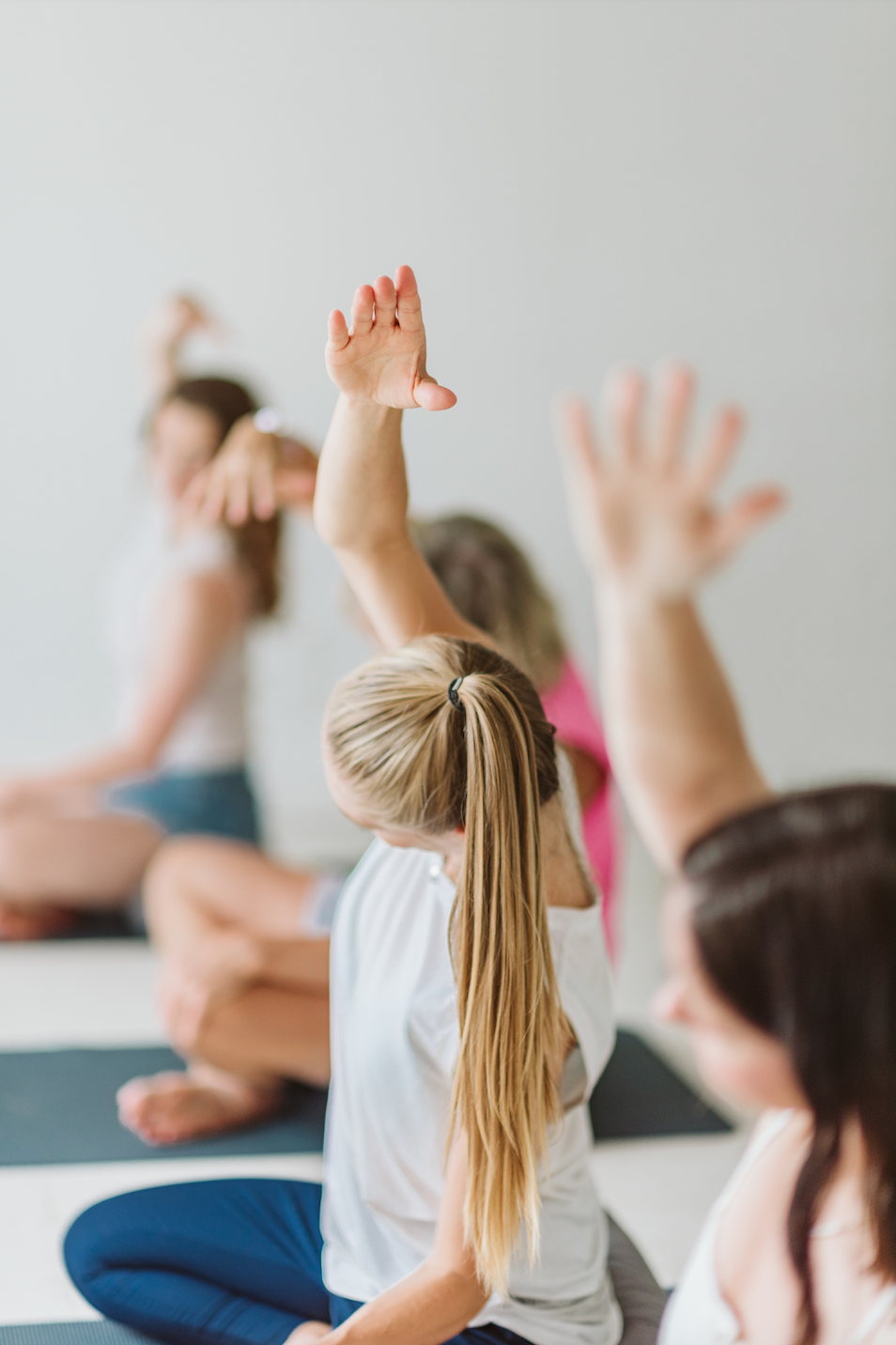 Children participating in a yoga or meditation class, with some raising their hands and others sitting cross-legged in a classroom setting.