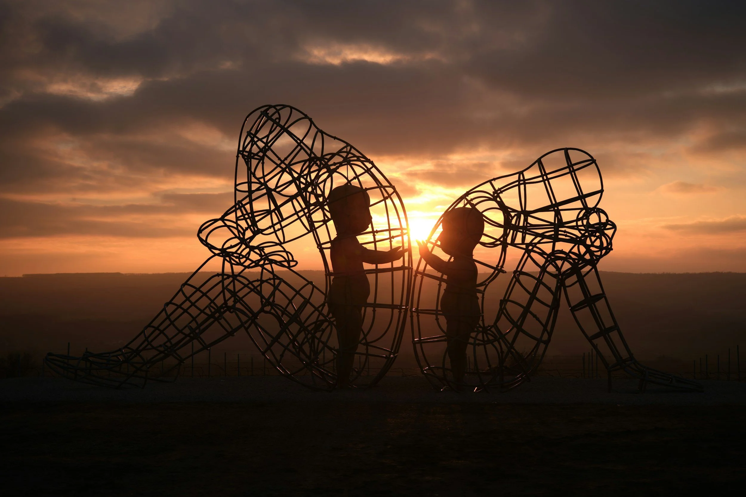Silhouette of two children touching hands inside a metal sculpture of a butterfly during sunset.