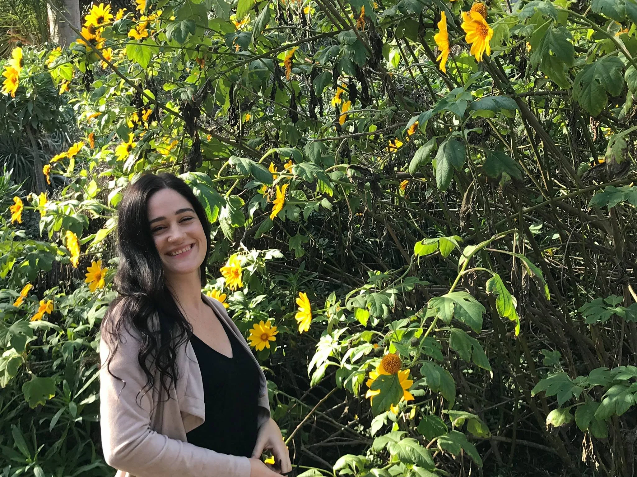 A woman with long dark hair smiling in a garden filled with yellow flowers.