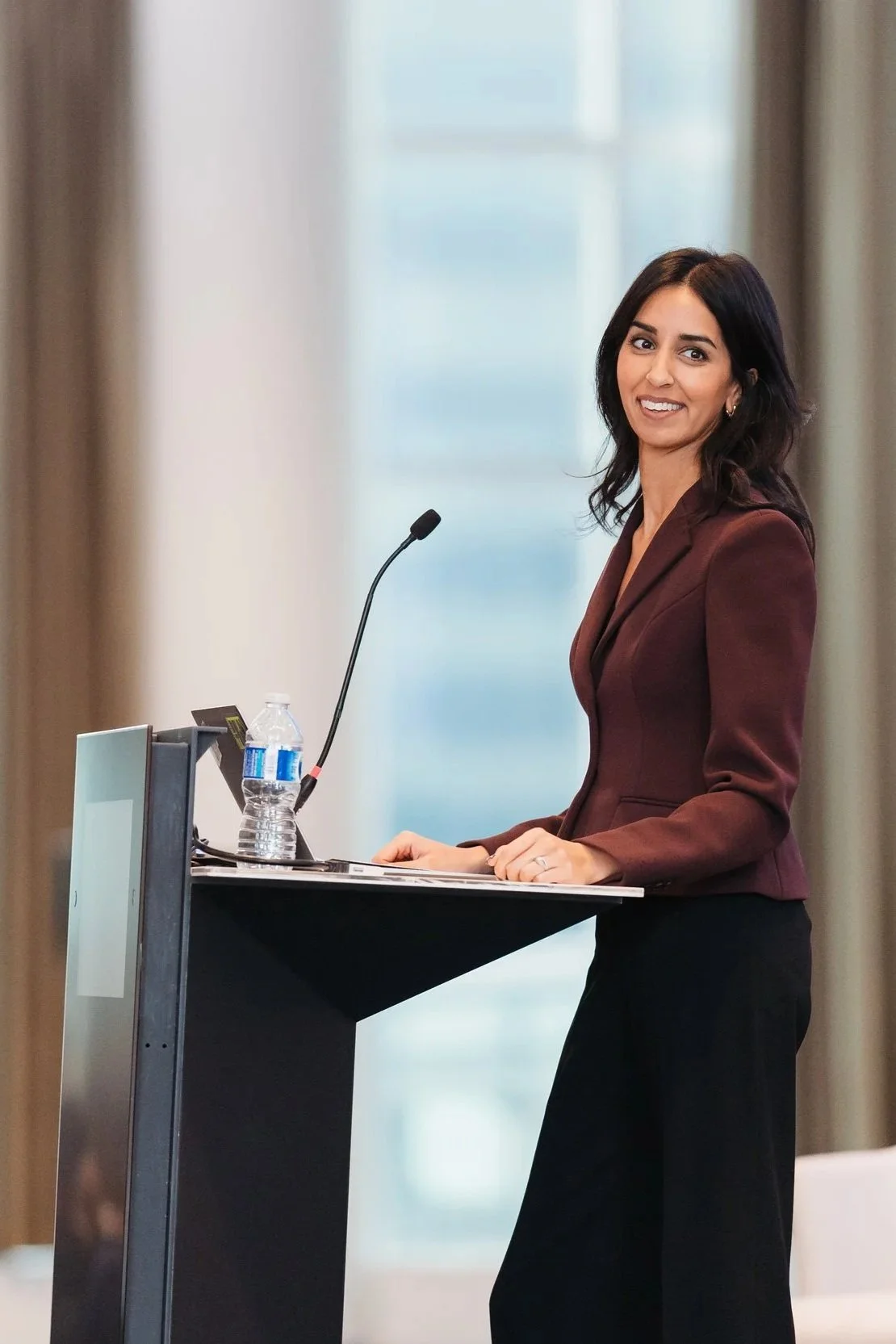 A woman with dark hair, wearing a maroon blazer, standing behind a podium and smiling at the camera during a speech or presentation.
