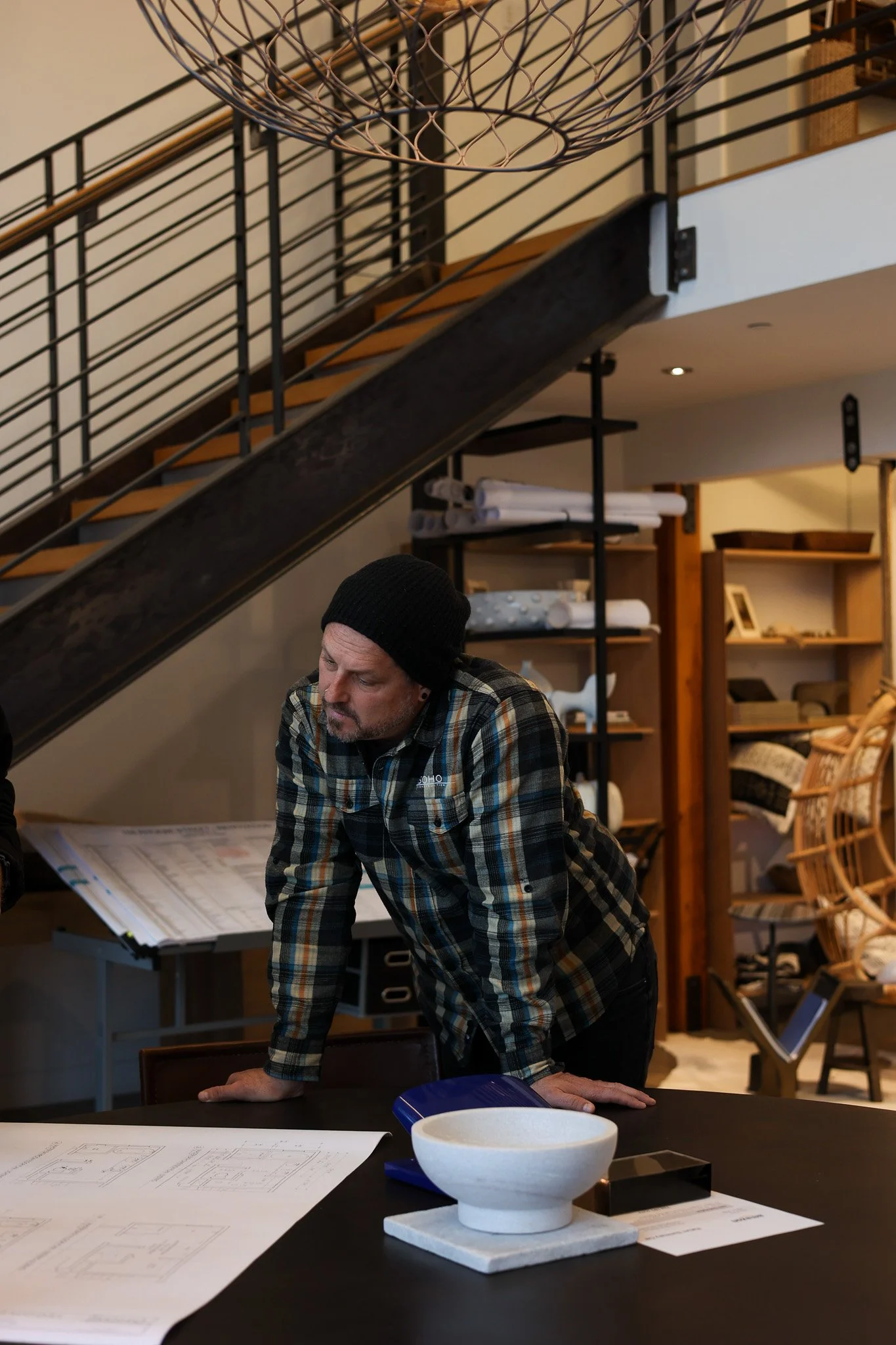 A man in a plaid shirt and black beanie leans forward on a table, looking at architectural plans in an interior space with wooden shelves, a staircase, and various home decor items.