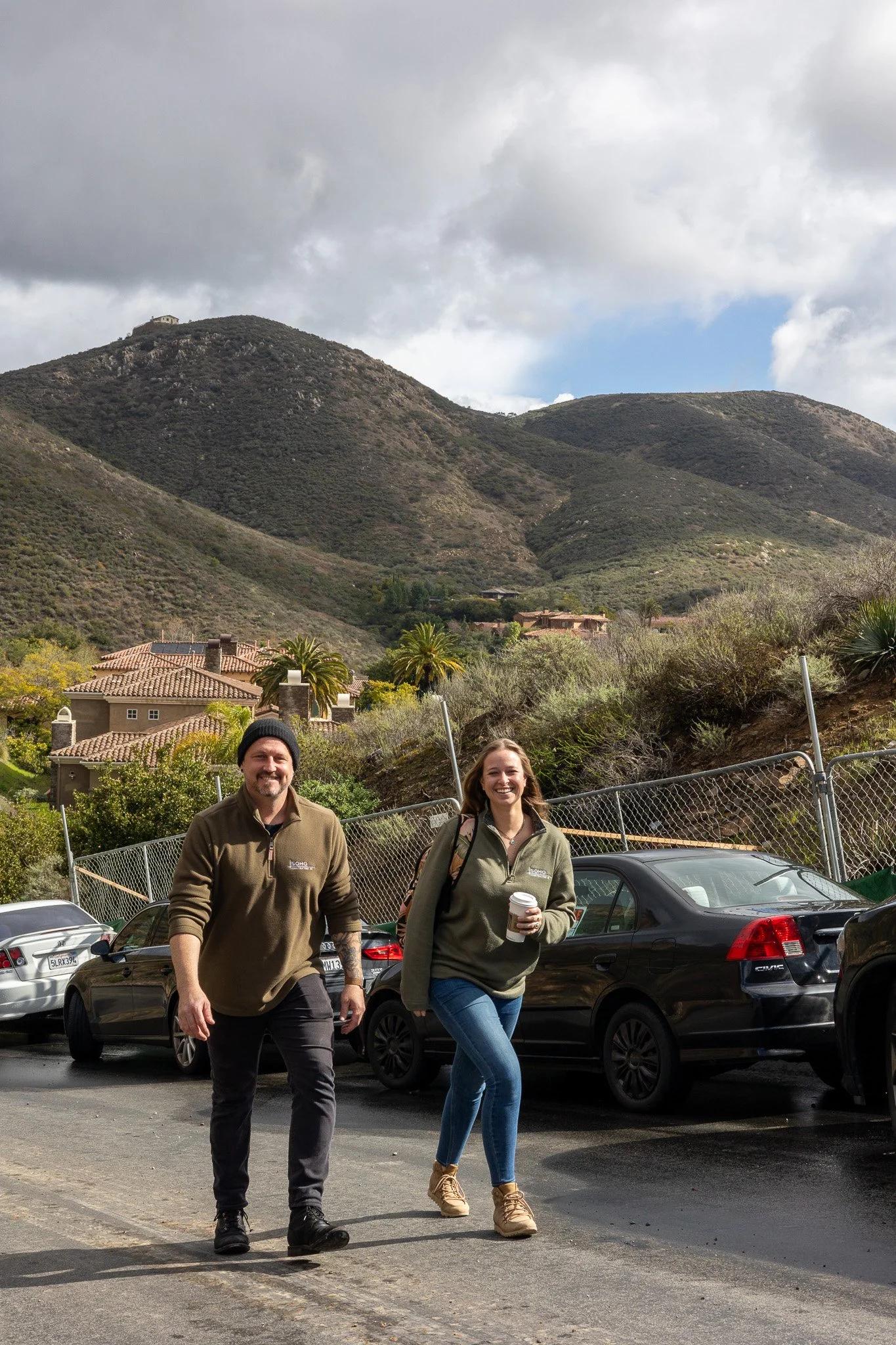 Two people walking in a parking lot with hills and residential houses in the background, one is holding a coffee cup.