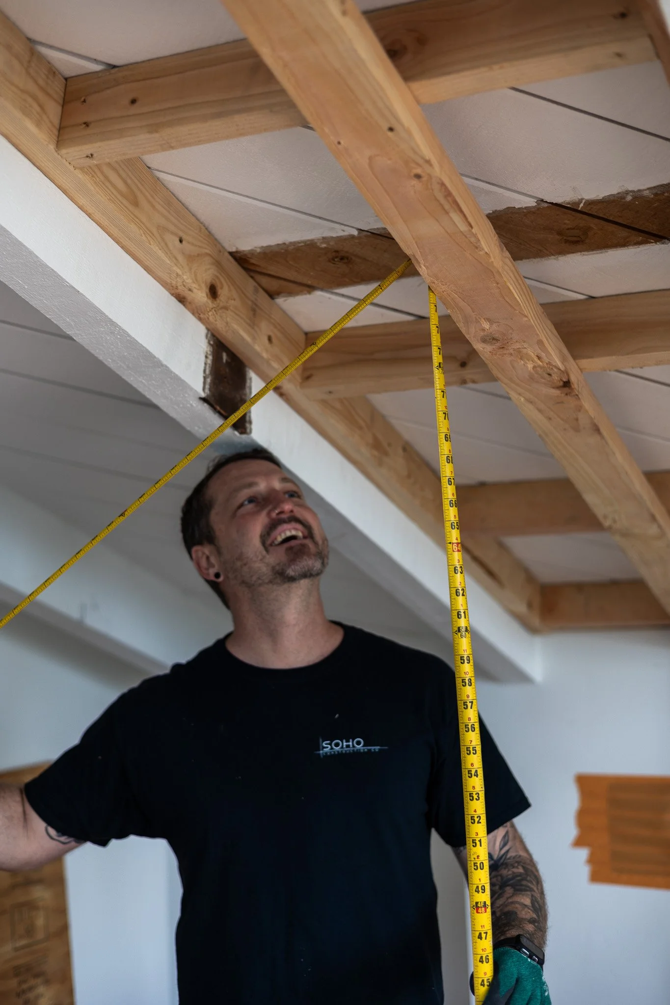 A man standing indoors under a partially constructed ceiling, measuring the height with a yellow measuring tape, smiling and looking up at the ceiling.