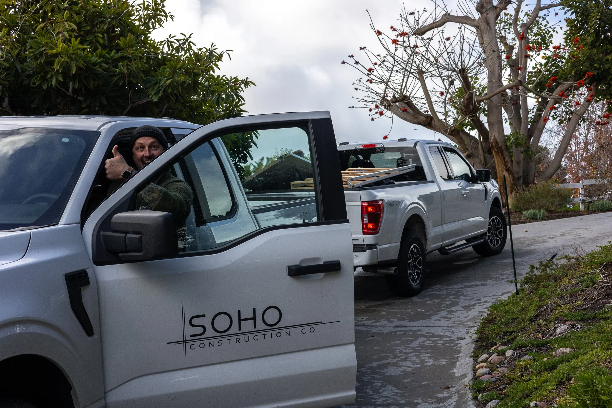 A smiling man in a dark beanie giving a thumbs-up while sitting in the driver’s seat of a white construction vehicle with 'SOHO CONSTRUCTION CO.' written on the door. Behind him, a white pickup truck is parked with wooden planks loaded in the bed. The scene is outdoors with trees and a cloudy sky visible in the background.