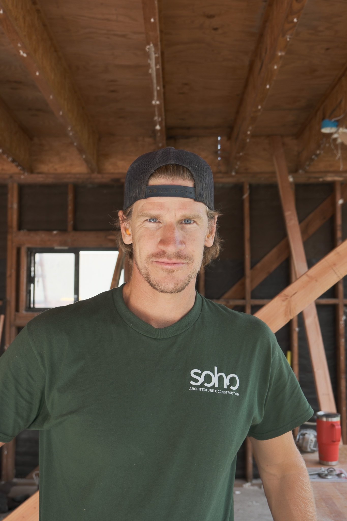 Two men are installing or repairing a ceiling or wall framework outdoors, with one standing on a ladder and the other holding a wooden beam, surrounded by construction tools and materials.