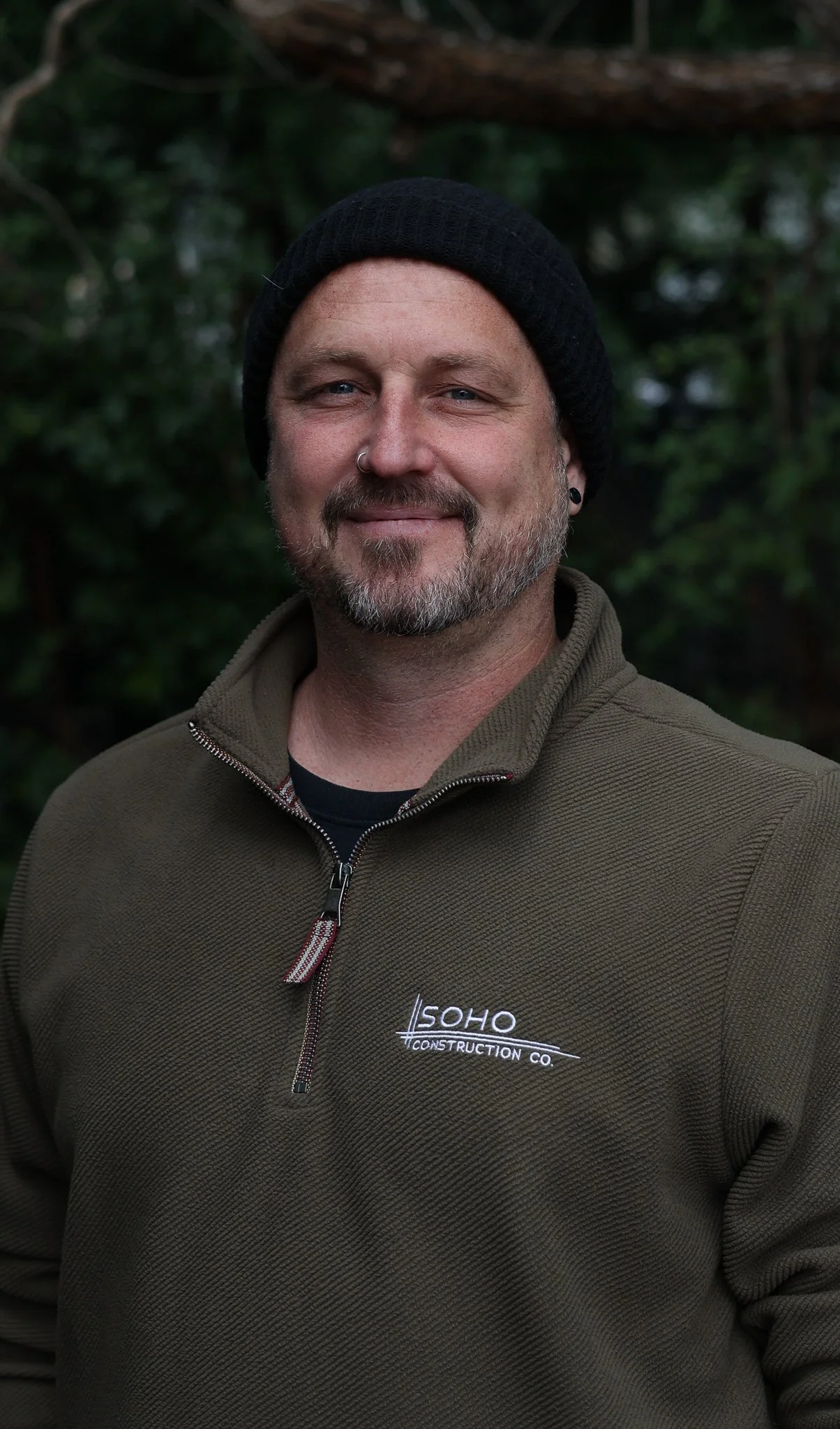 A smiling man with a beard and mustache wearing a black knit cap and a tan jacket with a zip, standing outdoors with blurred green foliage in the background.