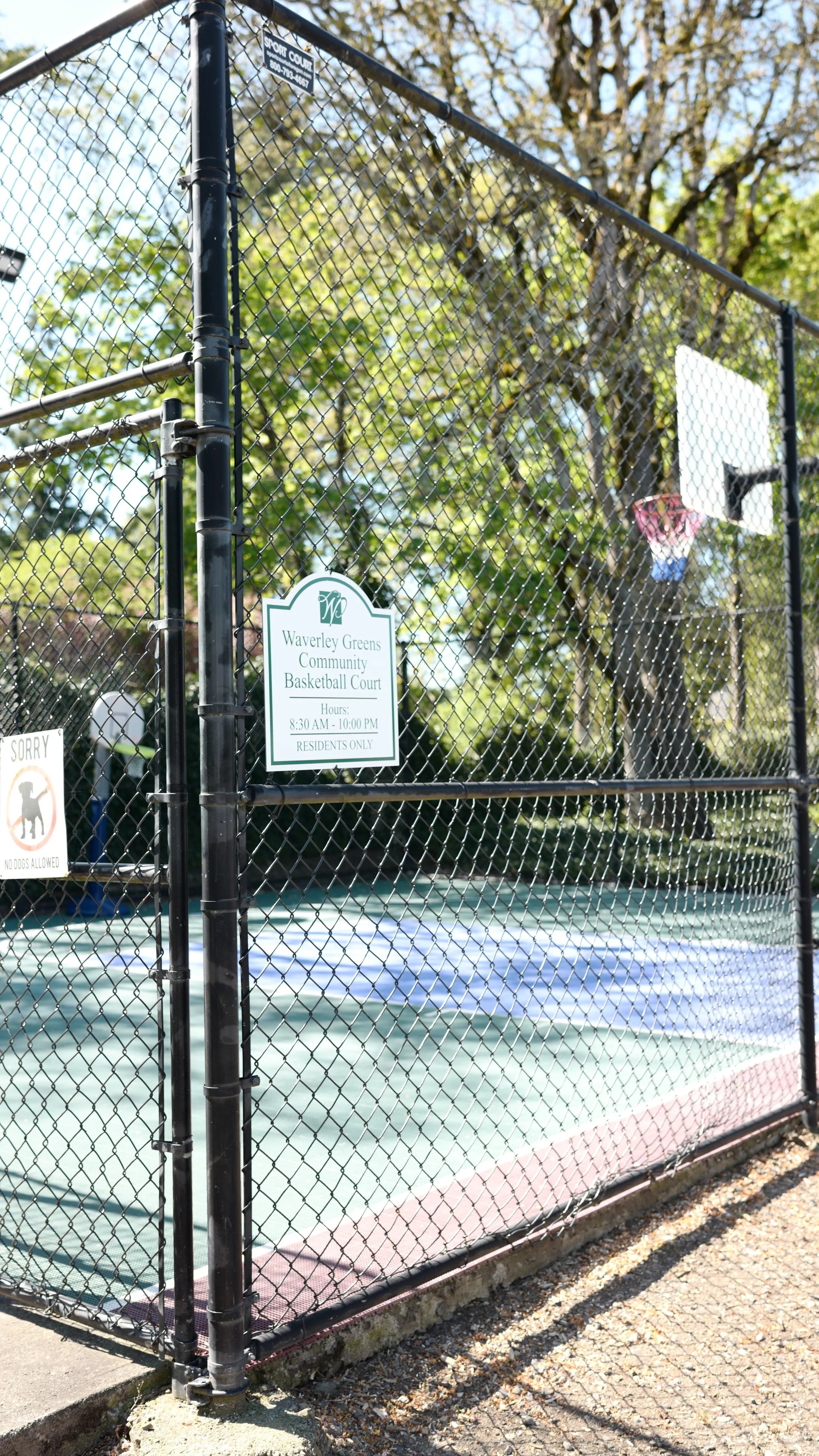 A fenced community basketball court with a sign displaying hours and rules, surrounded by trees on a sunny day.