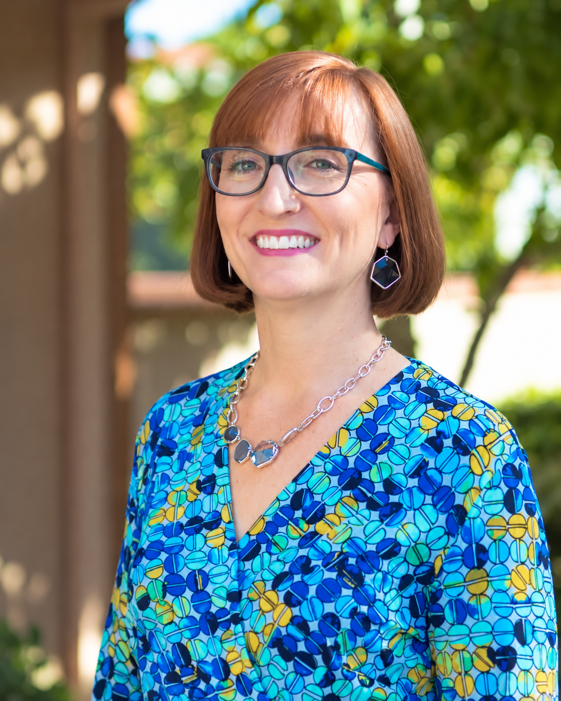 A smiling woman with short red hair, glasses, and earrings, standing outdoors with greenery in the background, wearing a colorful patterned blouse and jewelry.