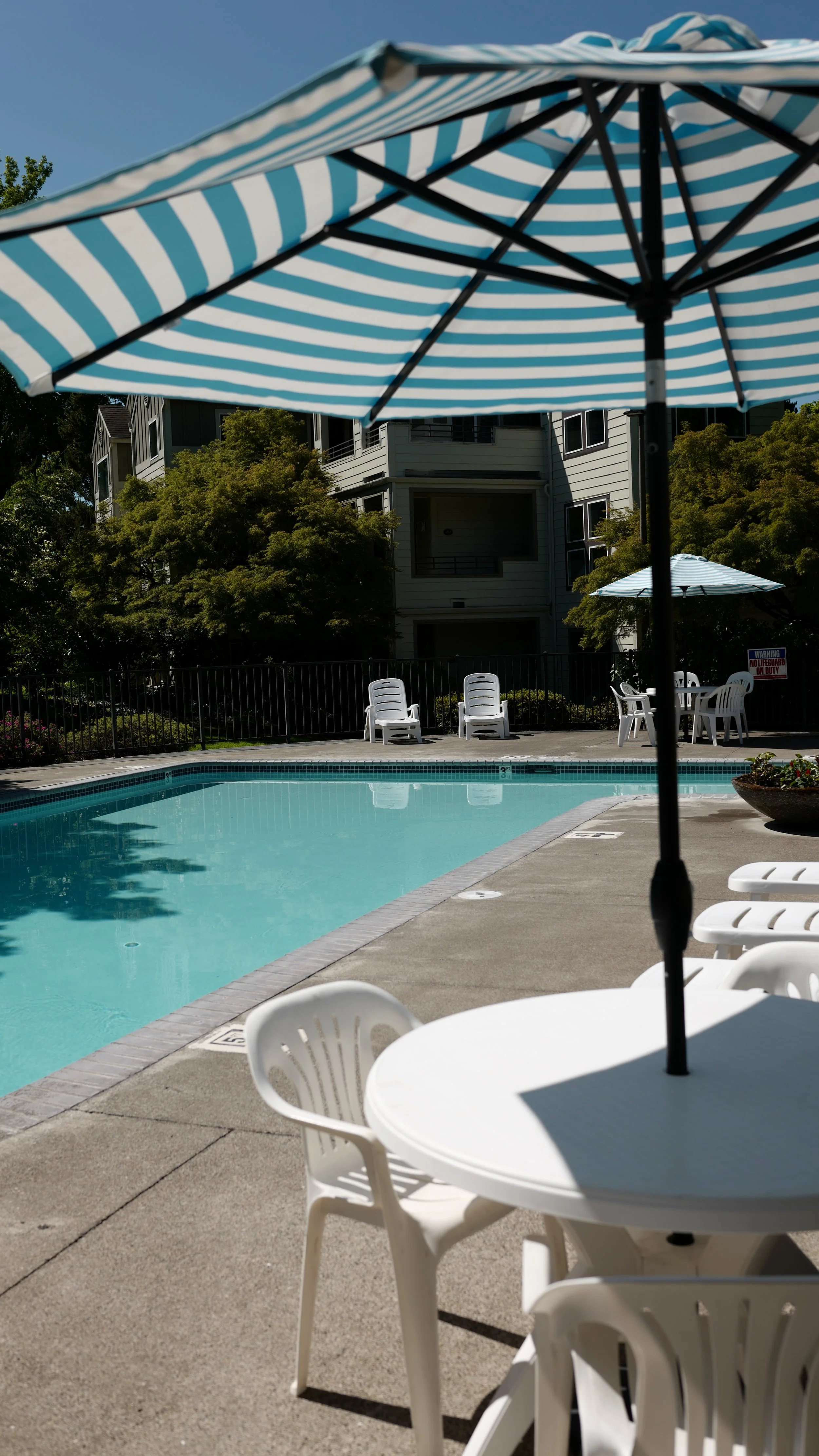 A swimming pool with a blue water surface, surrounded by white plastic chairs and tables with umbrellas providing shade, set in an outdoor area with trees and residential buildings in the background.