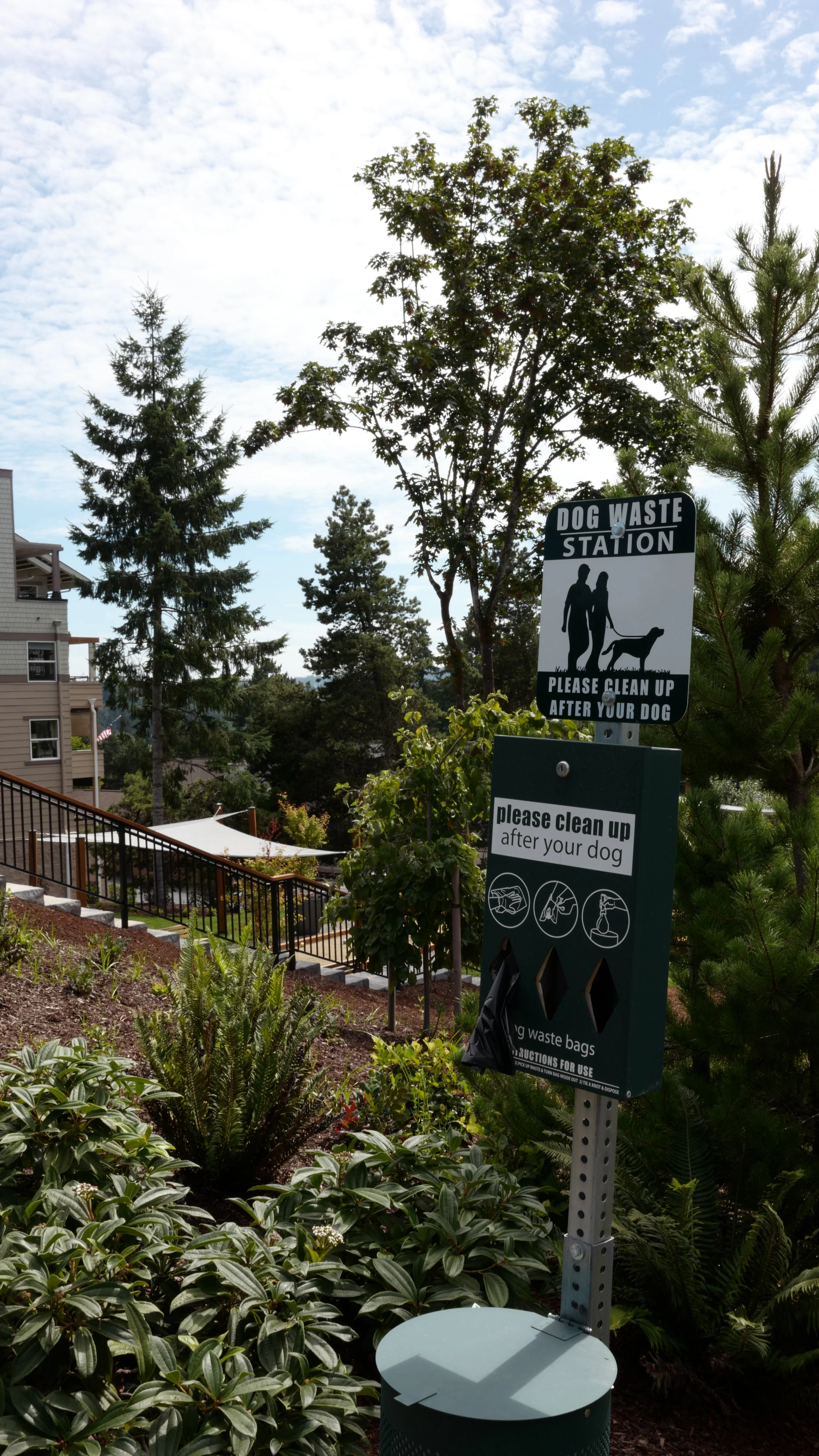 Dog waste station with a sign instructing pet owners to clean up after their dogs, surrounded by trees and plants in an outdoor area.