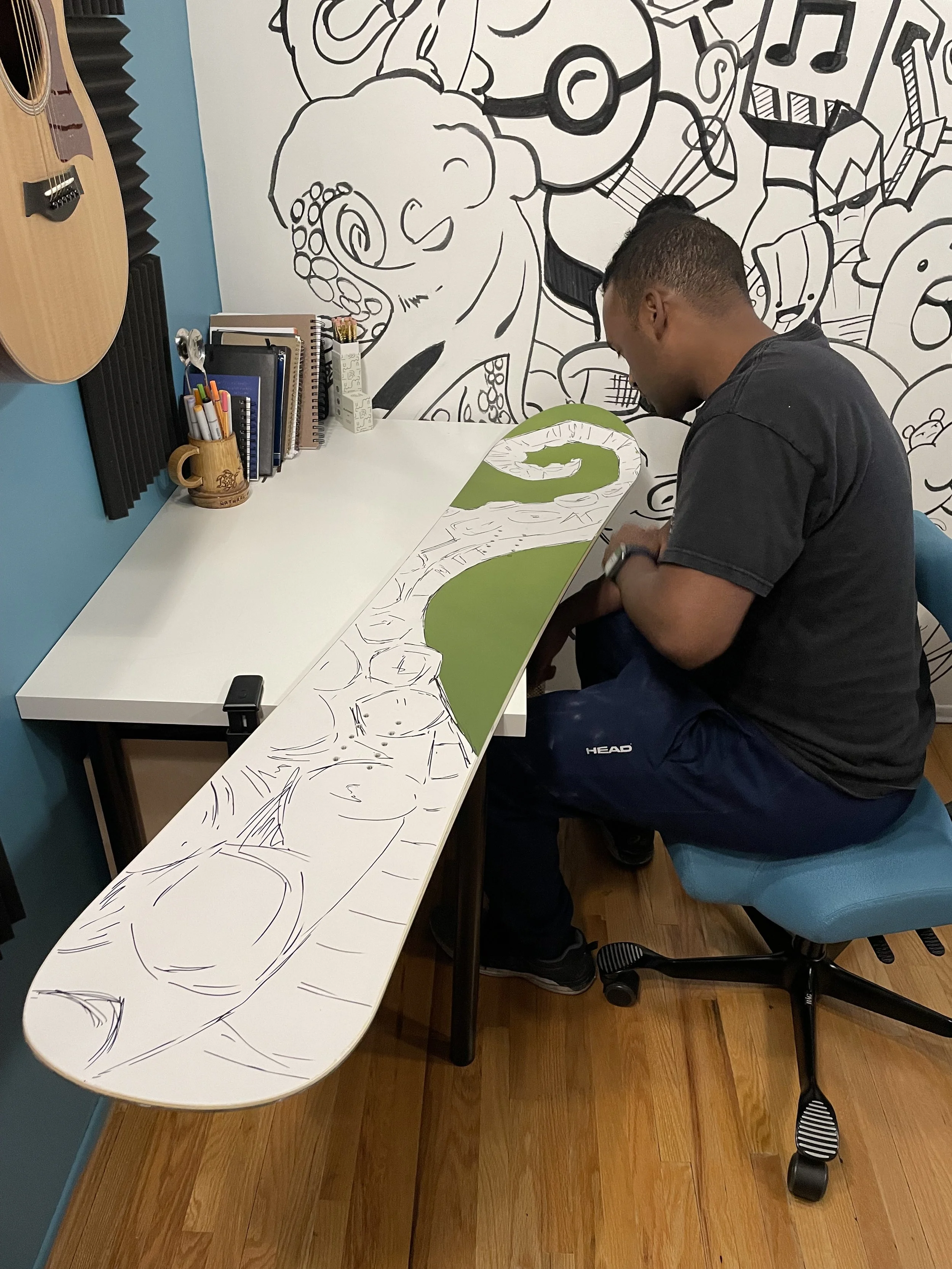 A person sitting in a blue office chair at a desk with a large sketch of cartoon characters on the surface, with a black and white wall drawing of cartoon characters in the background, and musical instruments on the wall.