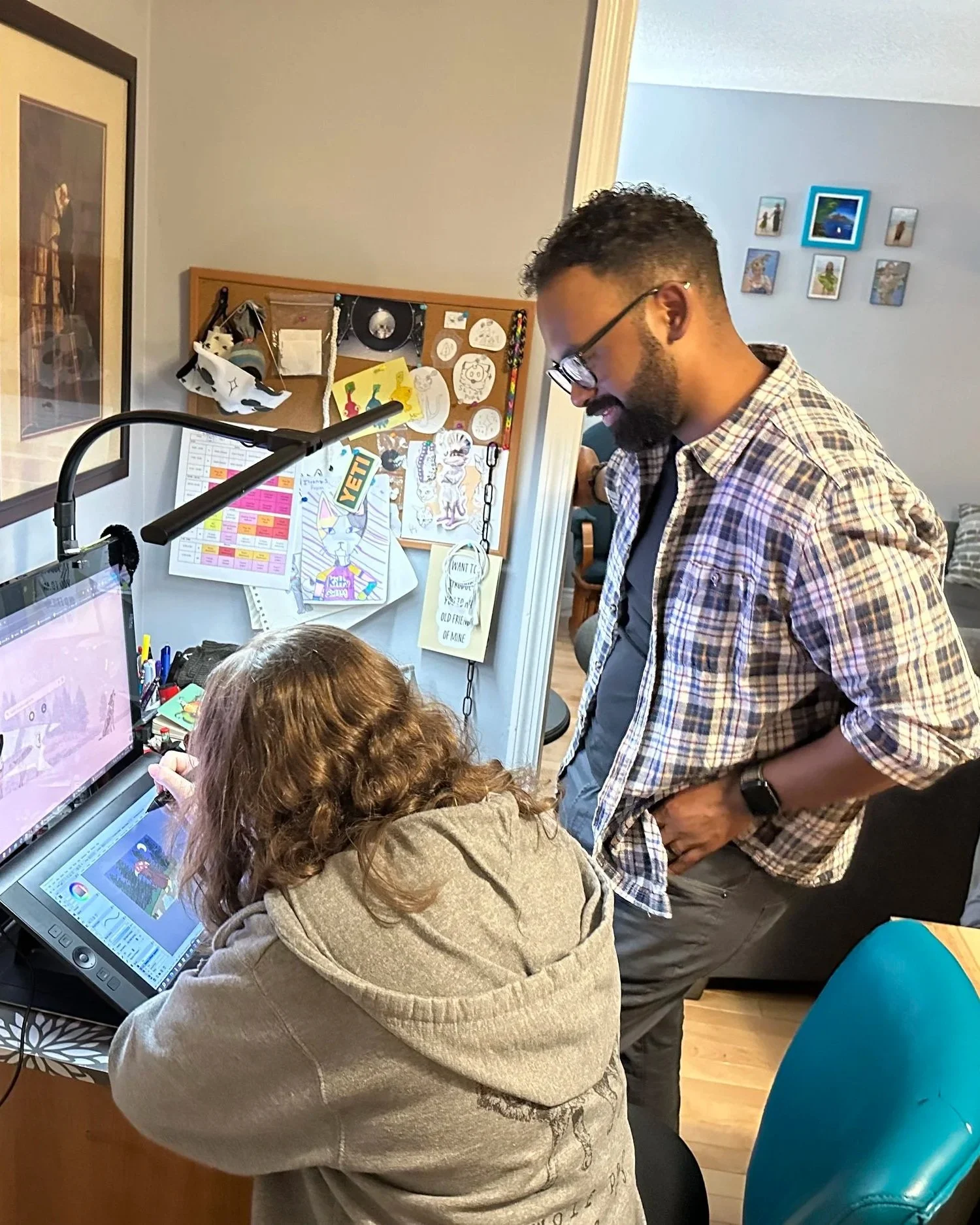 A young woman with curly red hair working on a digital drawing tablet, an older man with glasses standing beside her, both focused on the device. The background features a corkboard with various drawings and notes, a framed picture on the wall, and a small photo collage.