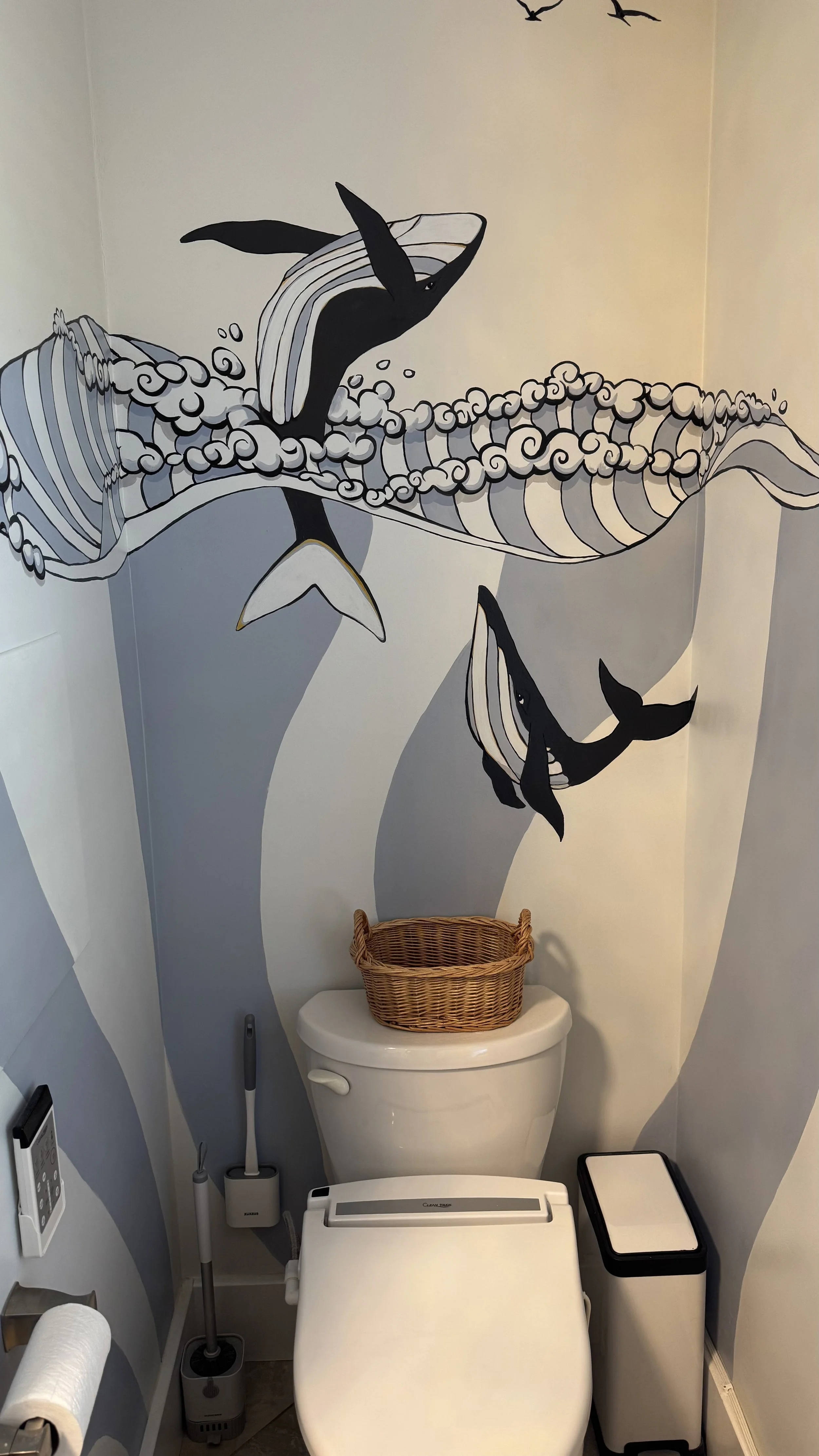 Bathroom with whale-themed watercolor mural on the wall behind the toilet. The mural depicts whales and ocean waves in black, white, gray, and blue tones. A wicker basket is placed on top of the toilet tank.