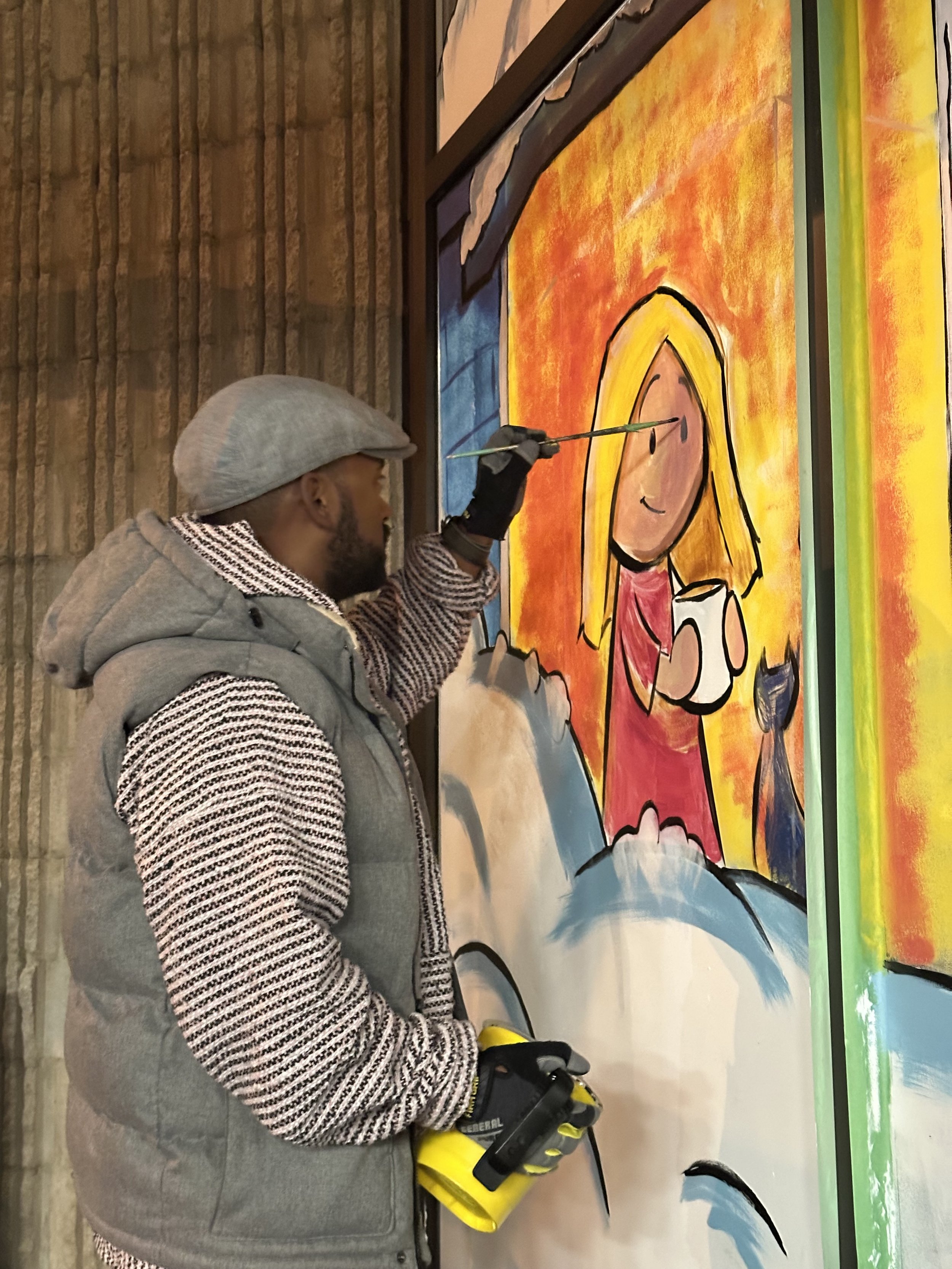 A person wearing a gray hat, black gloves, a striped sweater, and a gray vest that reads 'Federal' on the sleeve, is painting a colorful cartoon-style artwork on a large canvas or wall. The artwork depicts a smiling girl with long blonde hair, holding a cup, with a bright yellow and orange background. The person is holding a paintbrush.